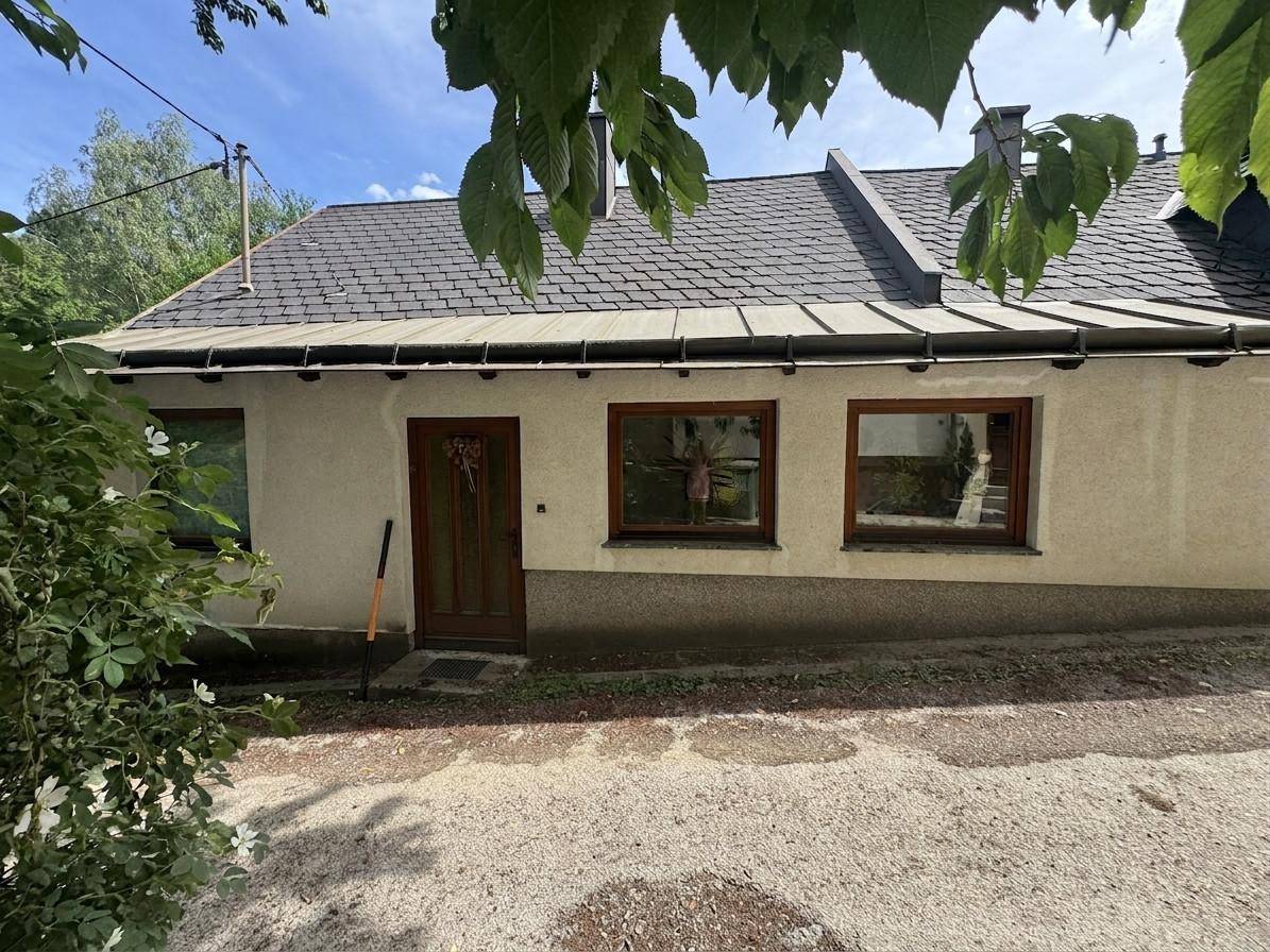 Vorderansicht eines Hauses mit Satteldach und braunen Fensterrahmen, umgeben von Bäumen.
