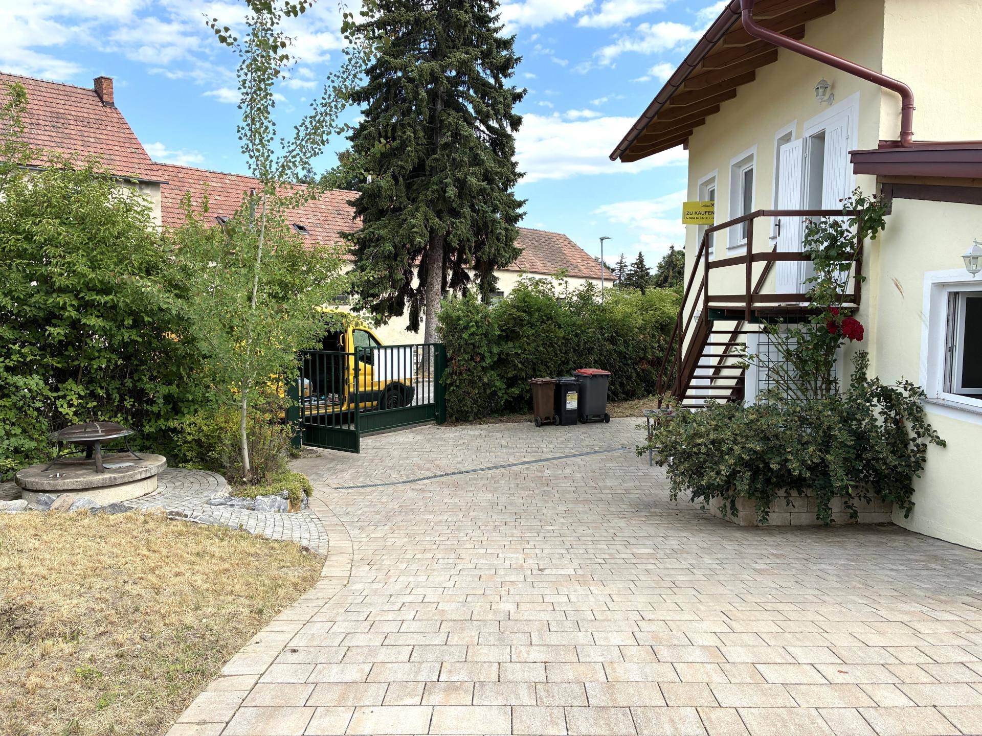 Gepflasterte Einfahrt mit grünem Tor und Bäumen neben dem Haus.