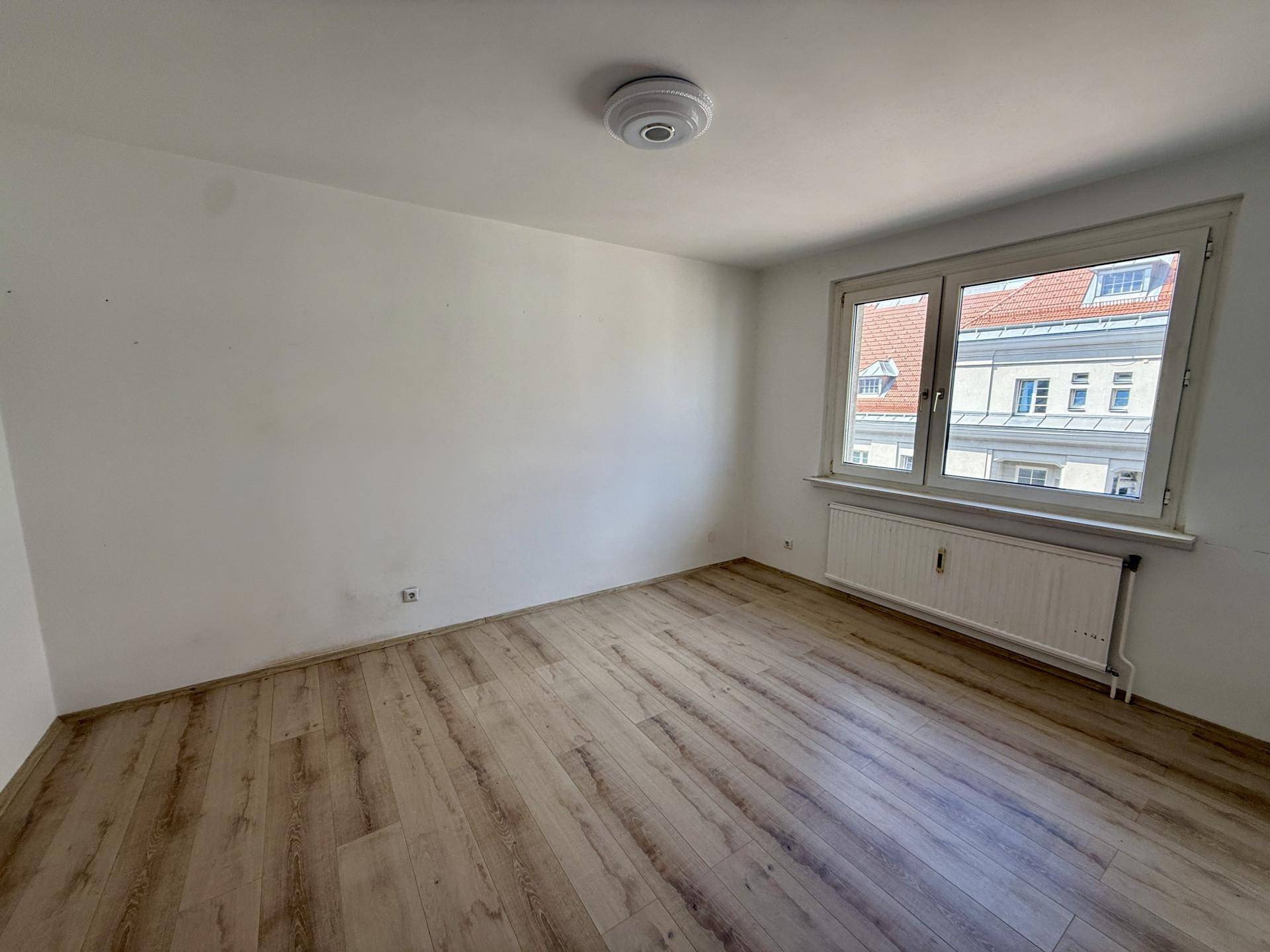 Leeres Zimmer mit Laminatboden, weißen Wänden und einem Fenster mit Blick auf Dächer.