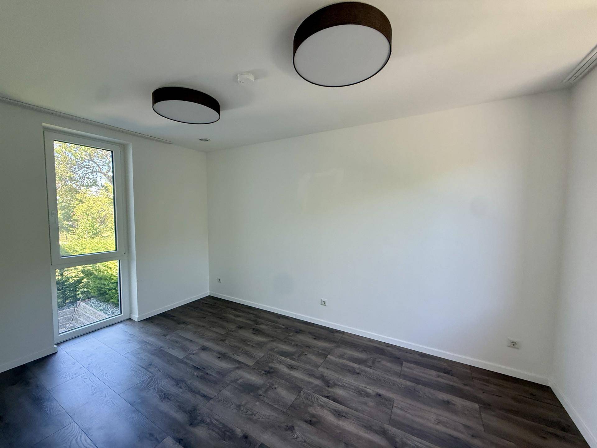 Helles Zimmer mit dunklem Laminatboden und großem Fenster mit Blick ins Grüne.