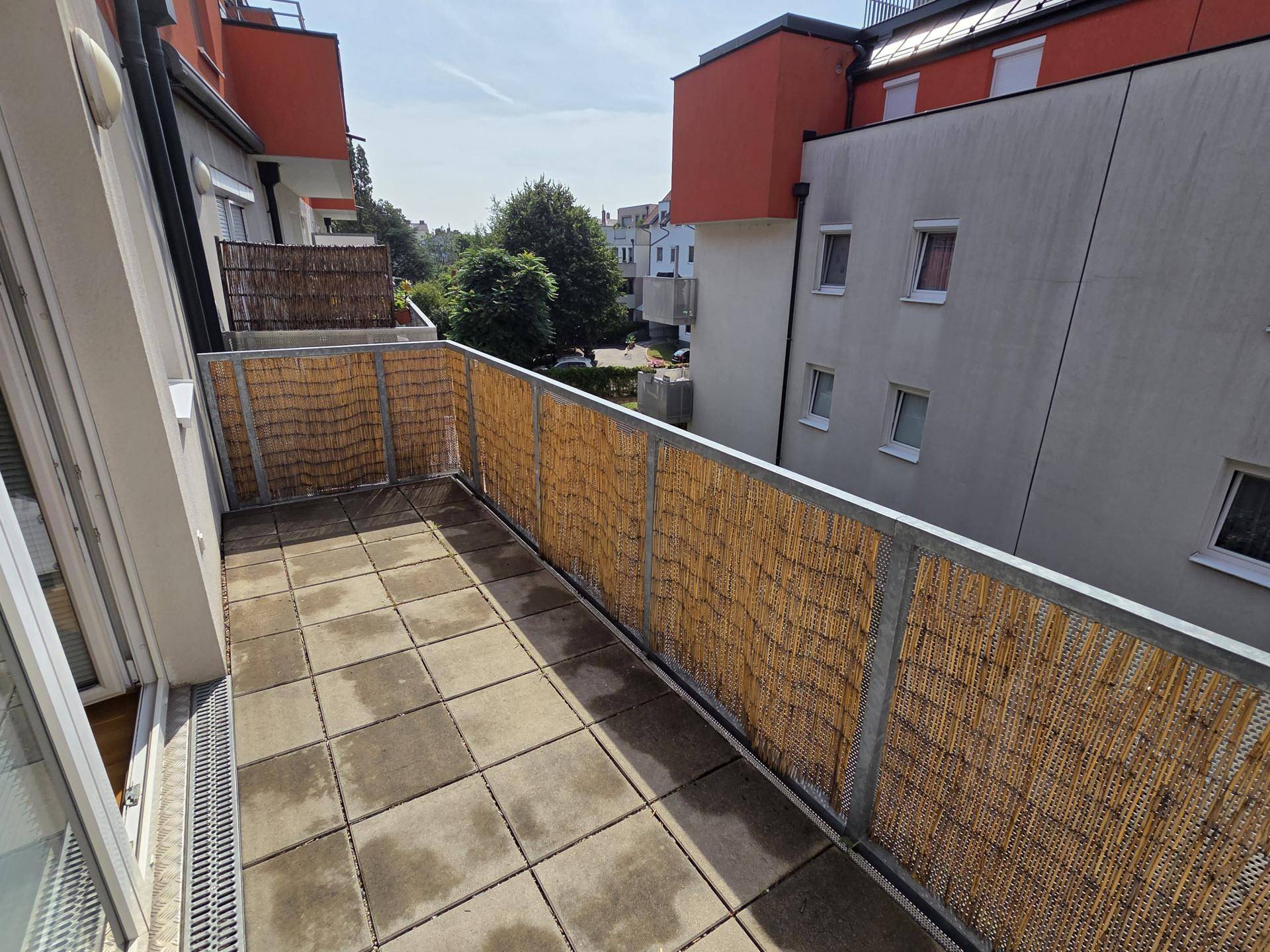 Langer Balkon mit Fliesenboden und Bambus-Sichtschutz, umgeben von Bäumen und Gebäuden.