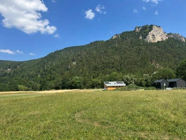 Weite grüne Wiese mit bewaldetem Berg und kleinen Gebäuden im Hintergrund unter blauem Himmel.