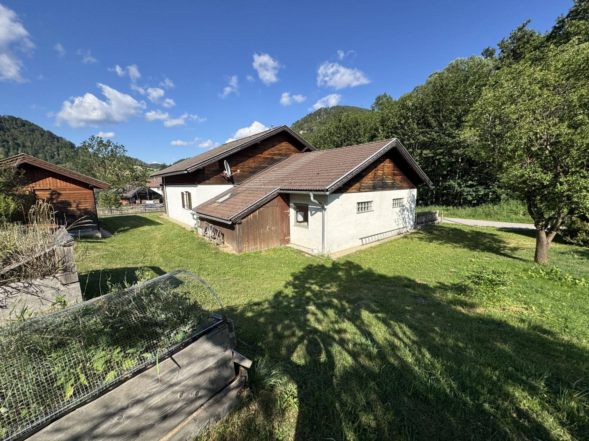 Haus mit Garten und Hochbeet in ländlicher Umgebung unter strahlend blauem Himmel.