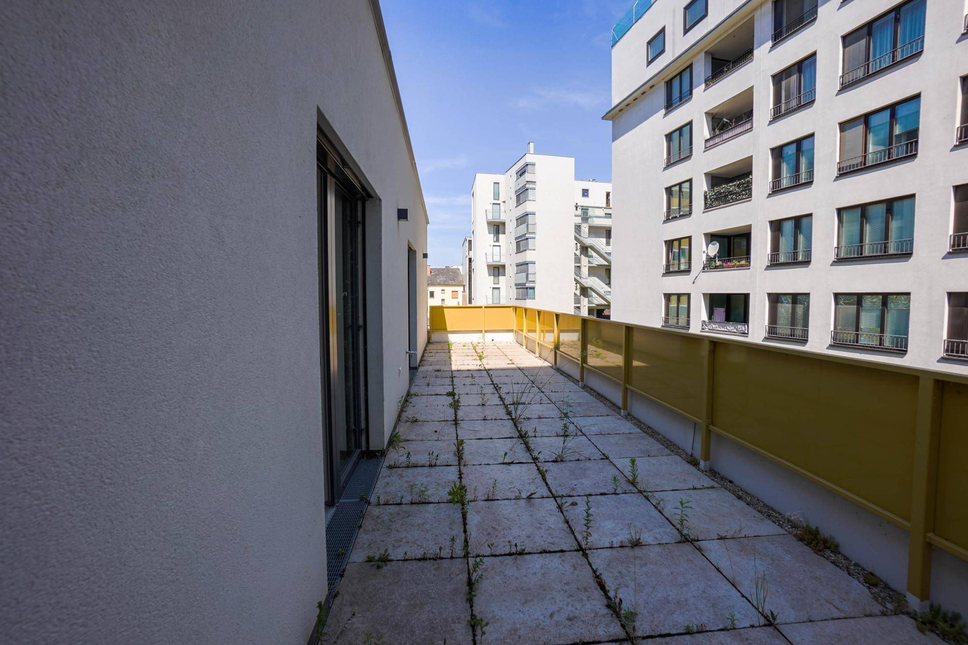 Lange, geflieste Terrasse mit gelbem Geländer und Blick auf moderne Wohngebäude.