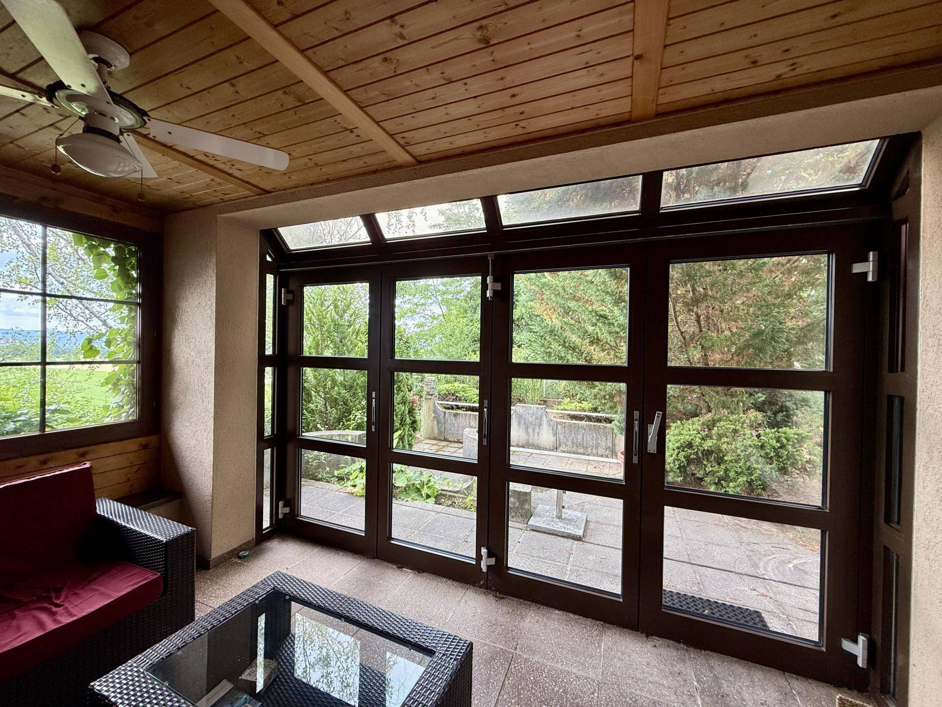 Überdachte Loggia mit dunklen Fensterrahmen, Fliesenboden und Deckenventilator, ideal für entspannte Stunden im Freien.