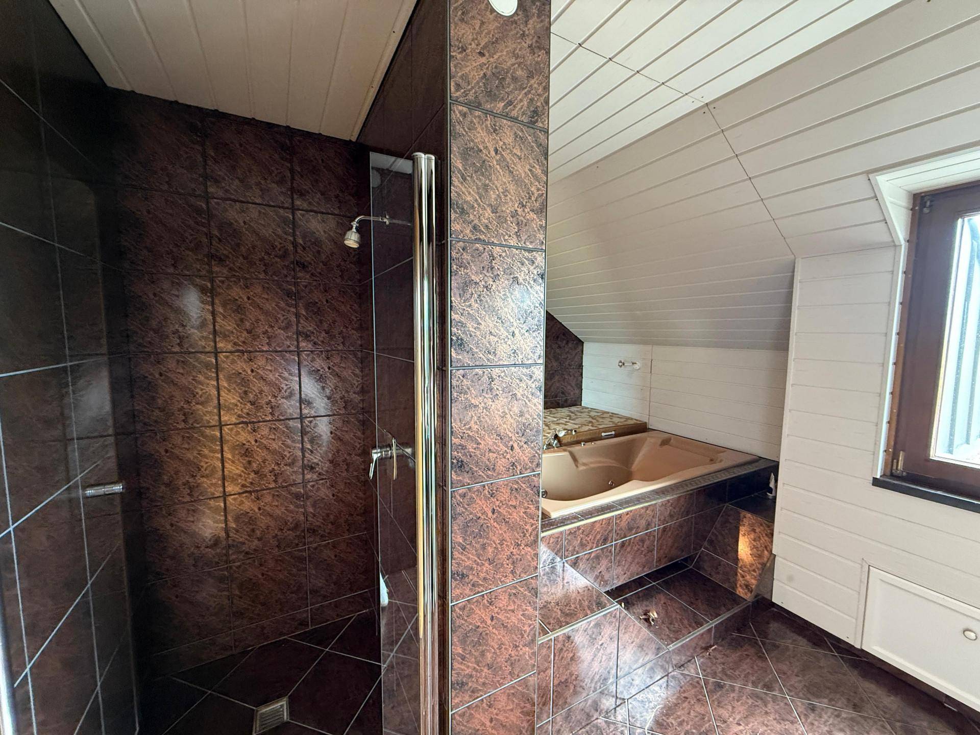 Badezimmer im Dachgeschoss mit braunen Fliesen, Dusche und Badewanne unter der Dachschräge, mit Fenster.