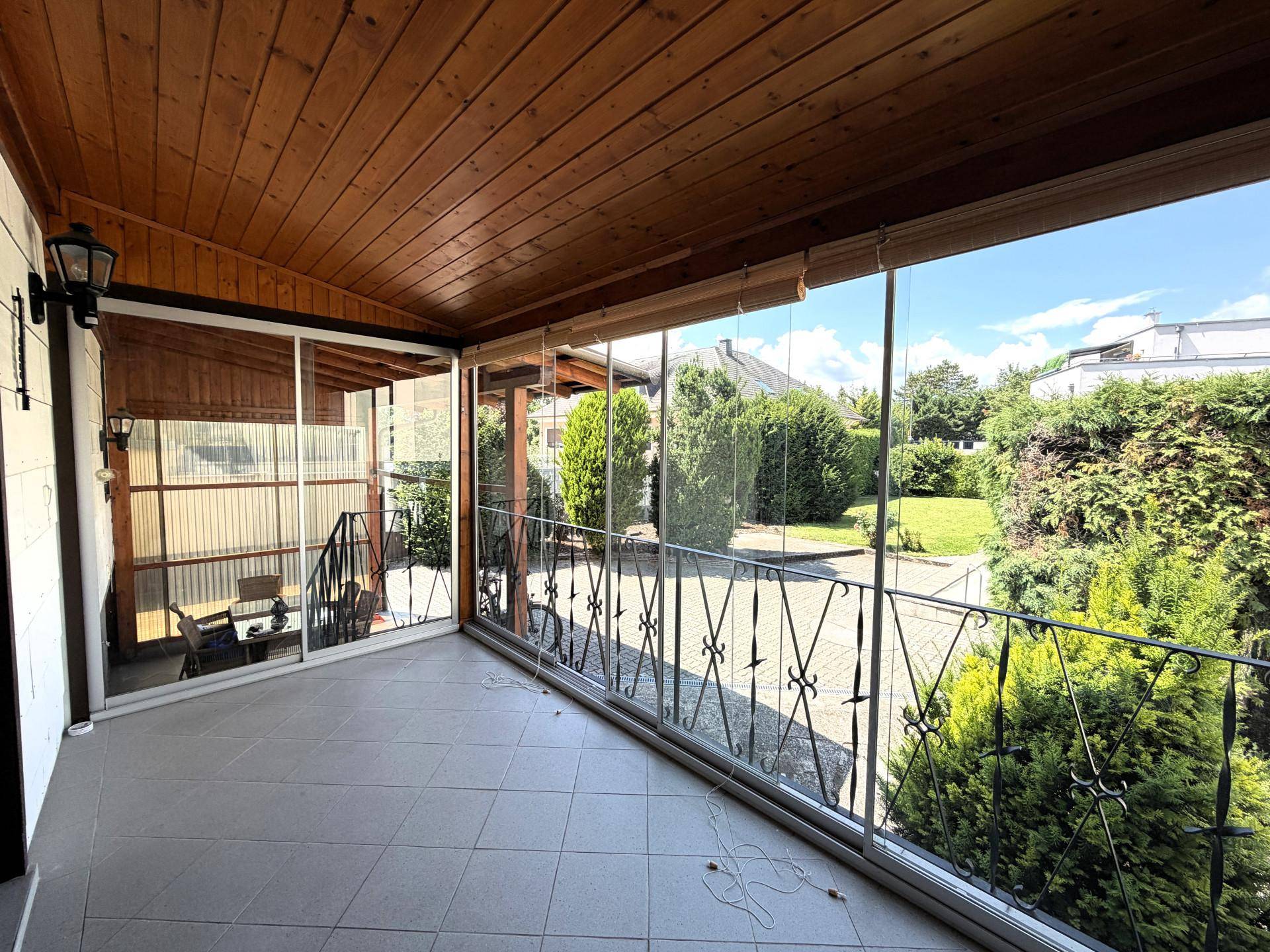 Verglaste Loggia mit Holzdach, Fliesenboden und Blick ins Grüne.