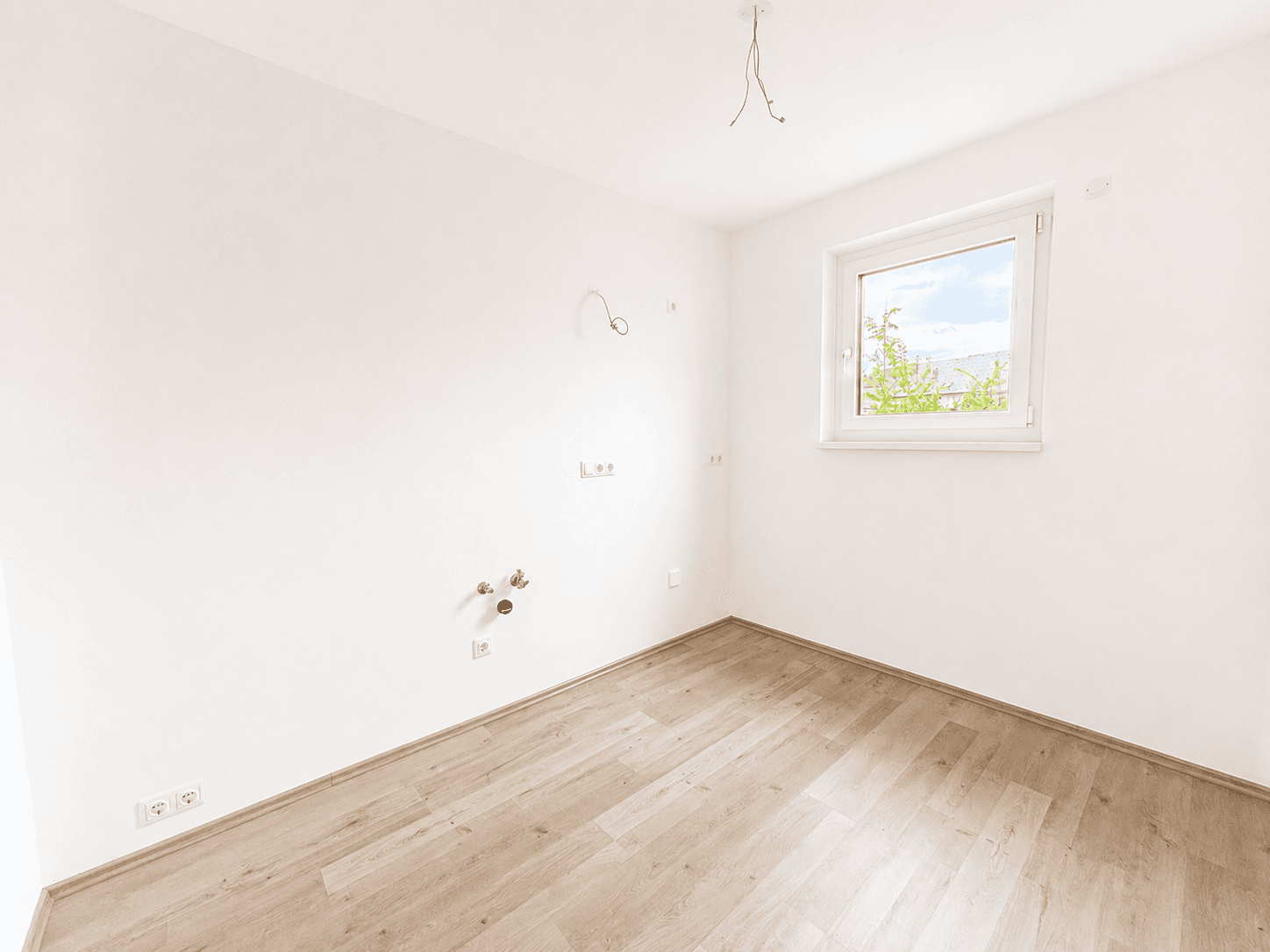 Leerer Hauswirtschaftsraum mit Anschlüssen für Geräte, Laminatboden und einem Fenster.