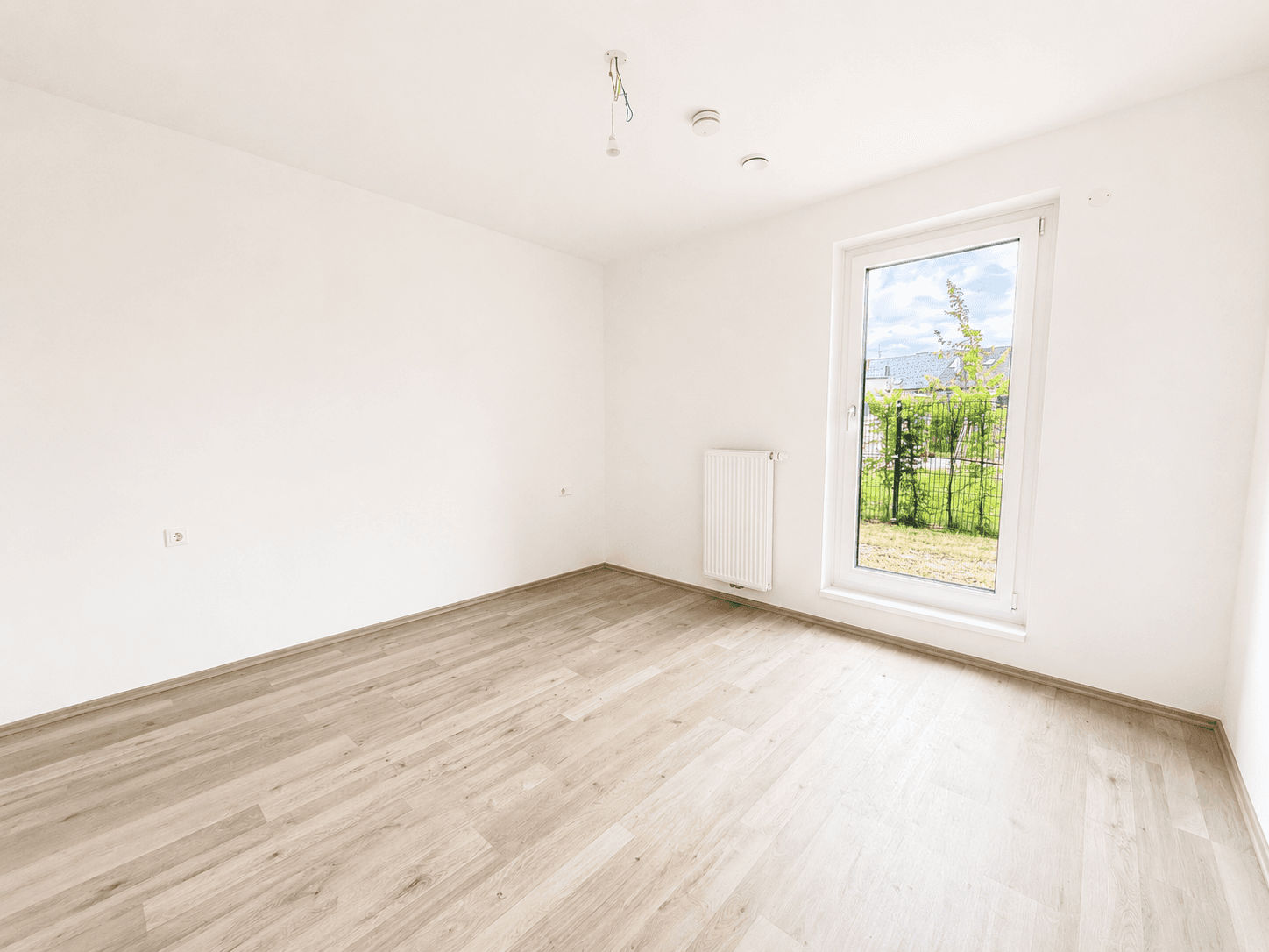 Leeres Schlafzimmer mit Laminatboden, Heizkörper und großem Fenster mit Blick ins Grüne.