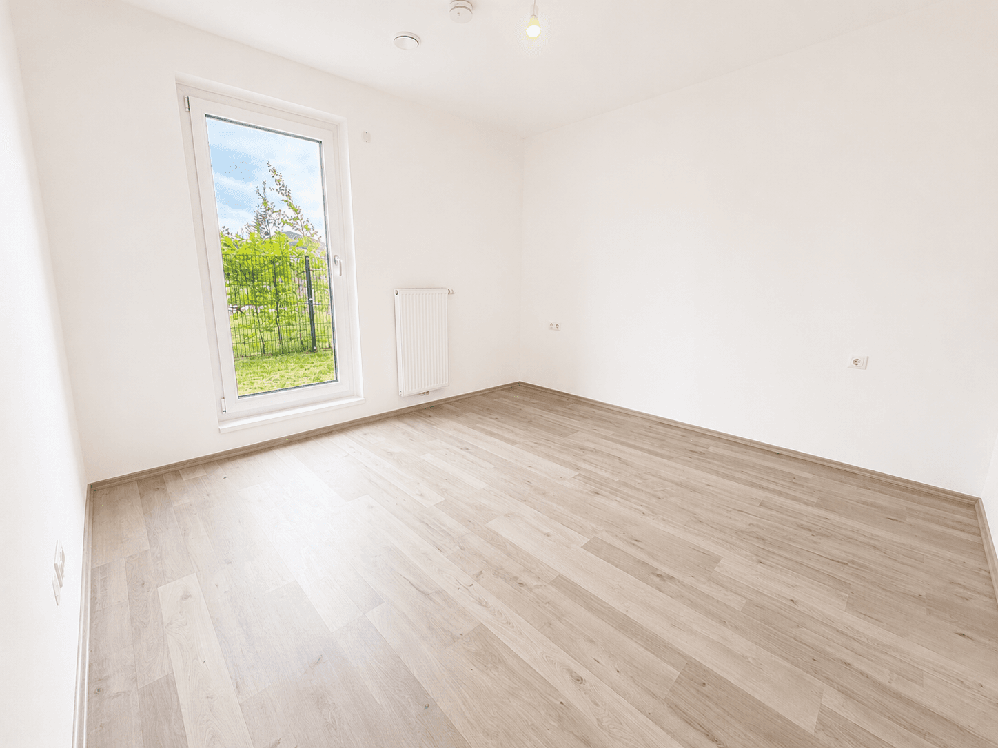 Leeres Schlafzimmer mit Laminatboden, weiß gestrichenen Wänden und Blick ins Grüne.