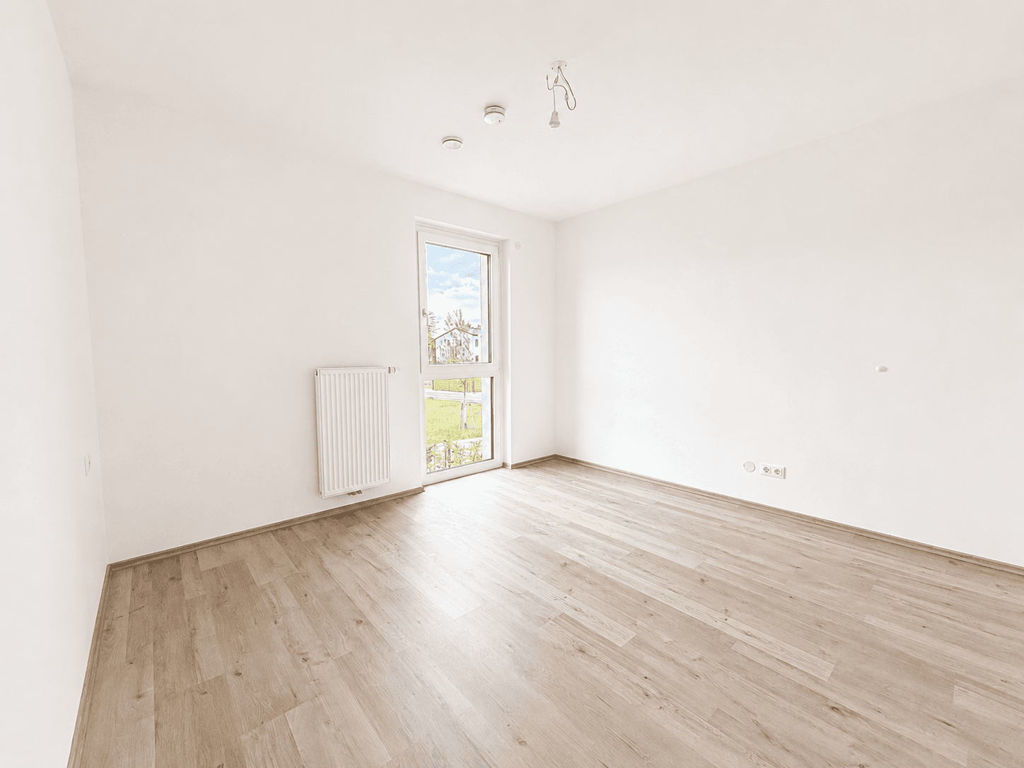 Leeres Schlafzimmer mit hellem Laminatboden, weißen Wänden und einem Fenster mit Blick ins Grüne.