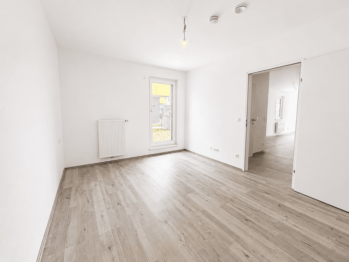Leerer, heller Raum mit Laminatboden, Fenster und Zugang zu einem weiteren Zimmer.