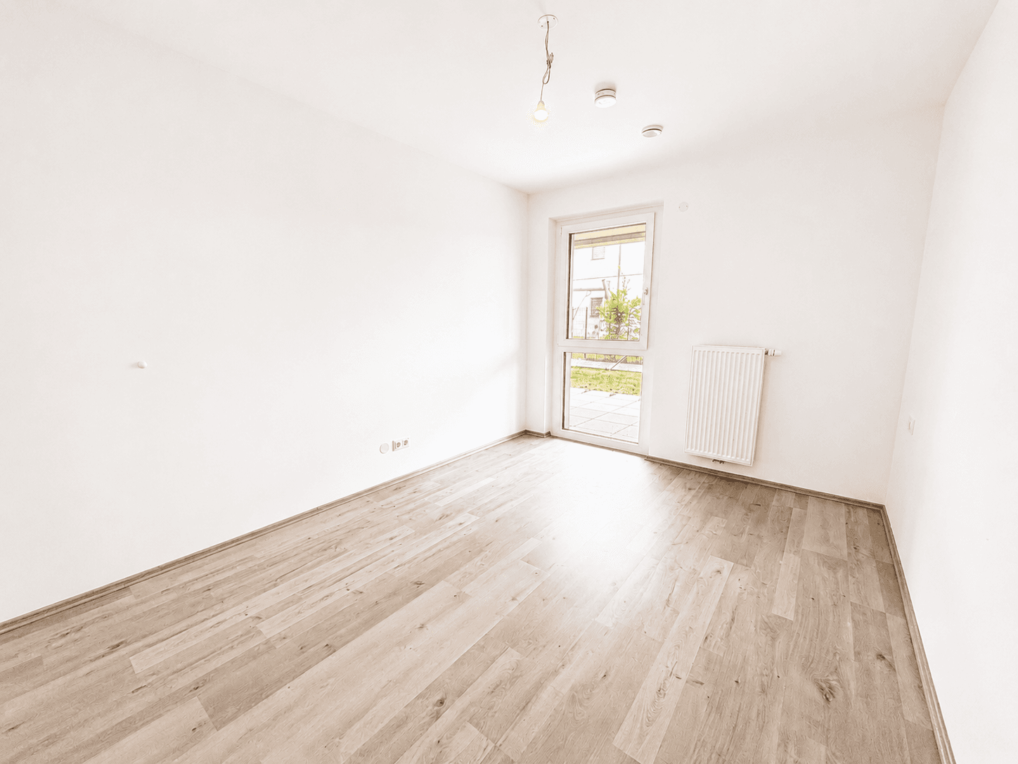 Leeres Schlafzimmer mit hellem Laminatboden, weißen Wänden und einem Fenster mit Zugang nach draußen.