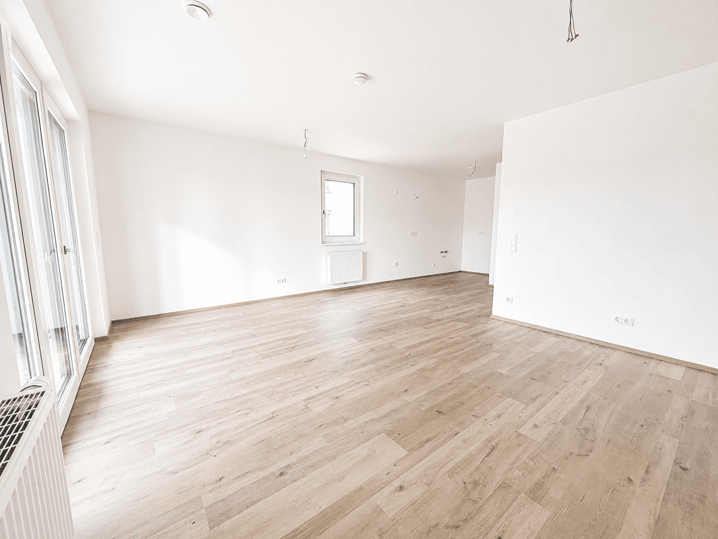 Geräumiges, unmöbliertes Wohnzimmer mit hellem Laminatboden und großen Fenstern.