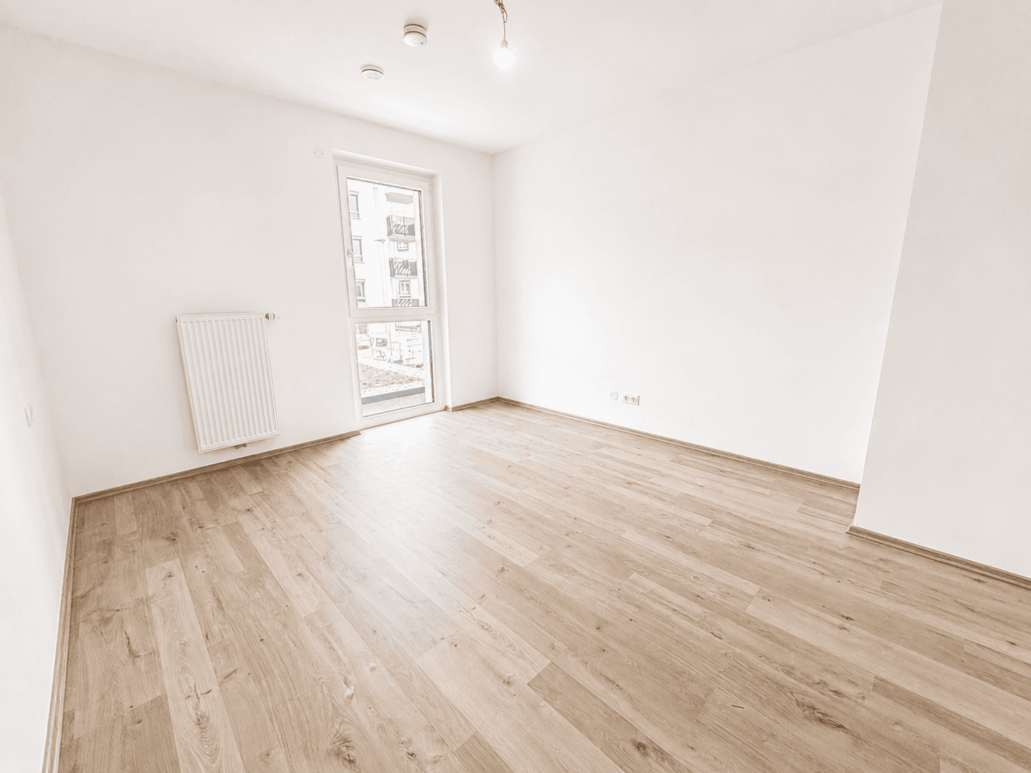 Leeres Schlafzimmer mit Laminatboden, Heizkörper und Fenster mit Blick nach draußen.