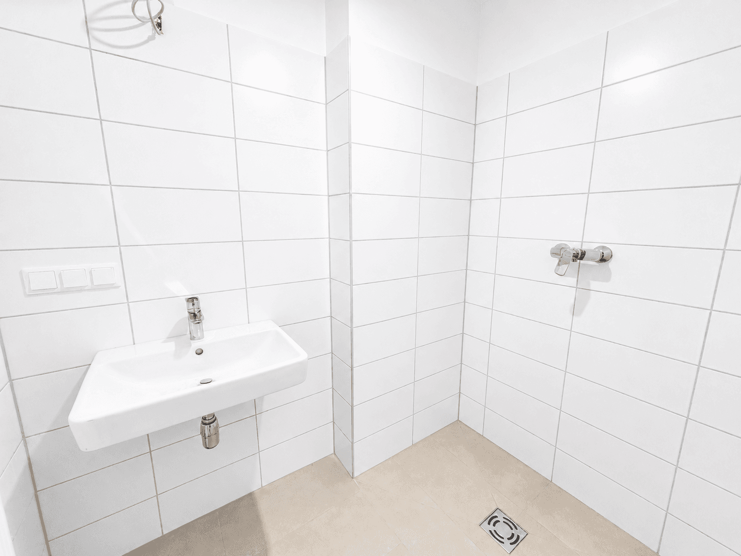 Helles Badezimmer mit weißen Wandfliesen, Waschbecken und ebenerdiger Dusche.