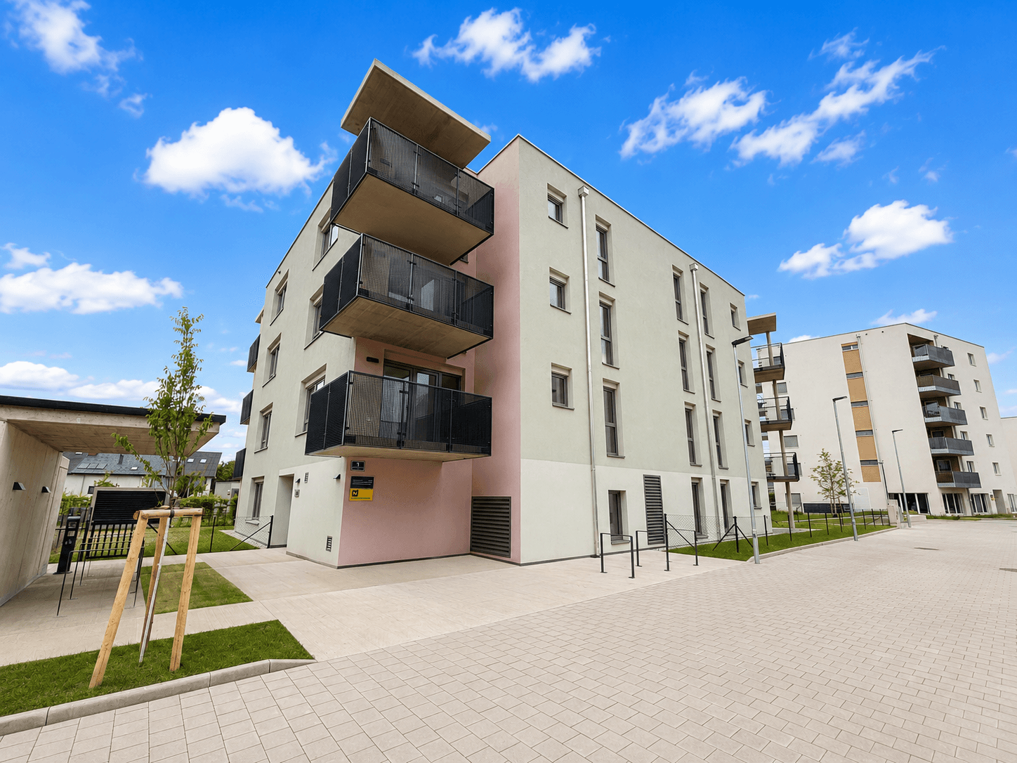 Modernes Wohngebäude mit Balkonen, heller Fassade und gepflastertem Vorplatz unter blauem Himmel.