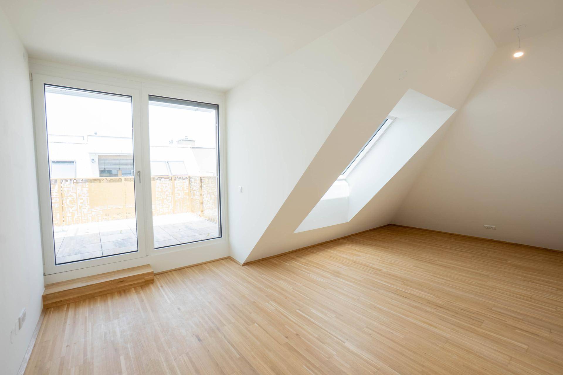 Heller Raum mit Parkettboden, schrägen Wänden und Zugang zu einem Balkon.