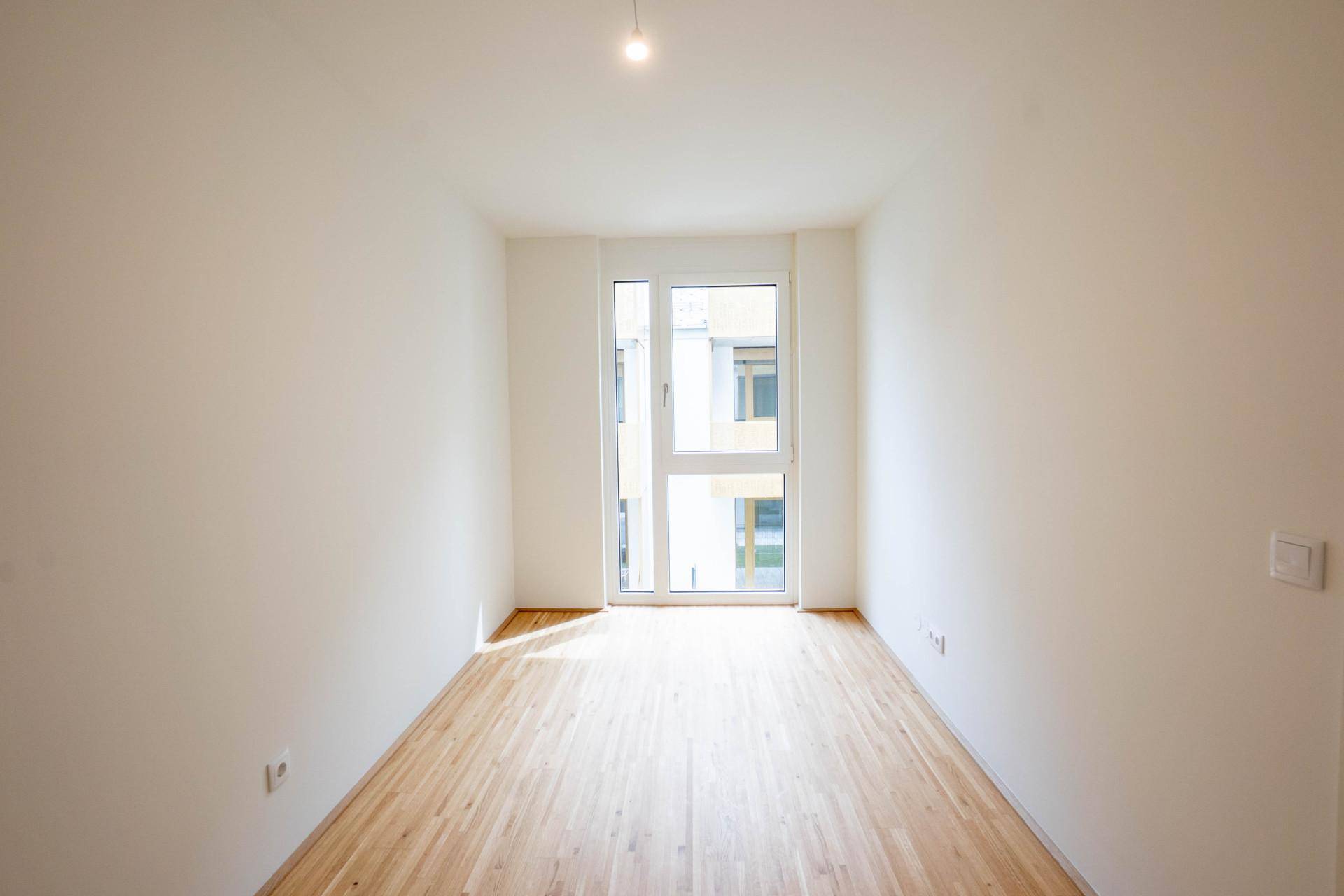 Ein helles, leeres Zimmer mit Holzboden und einem großen Fenster, ideal als Schlafzimmer.