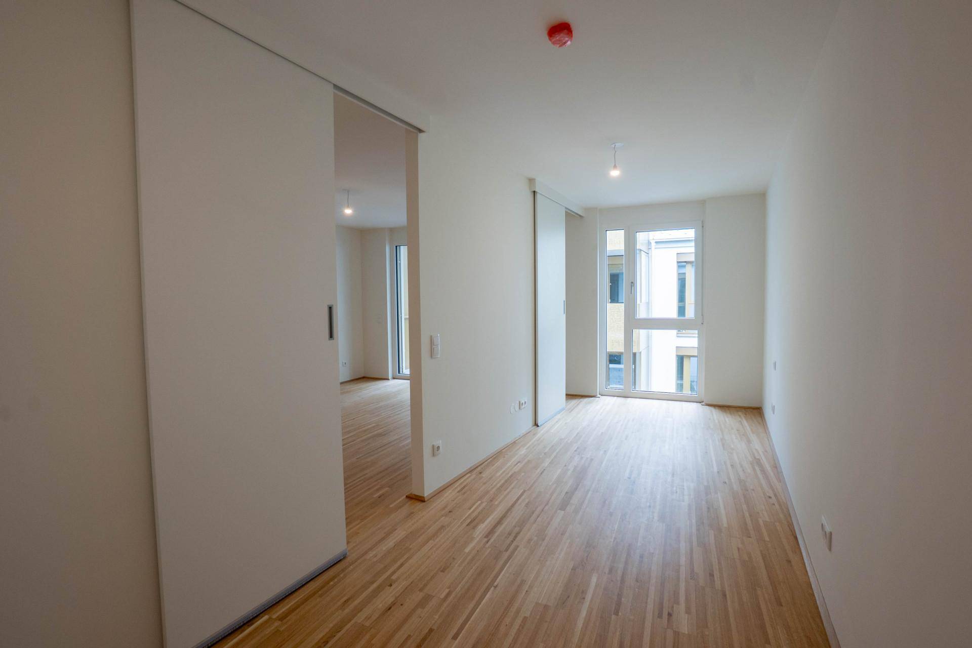 Heller Raum mit Holzboden und Schiebetüren, der zu weiteren Zimmern führt, mit viel Tageslicht.