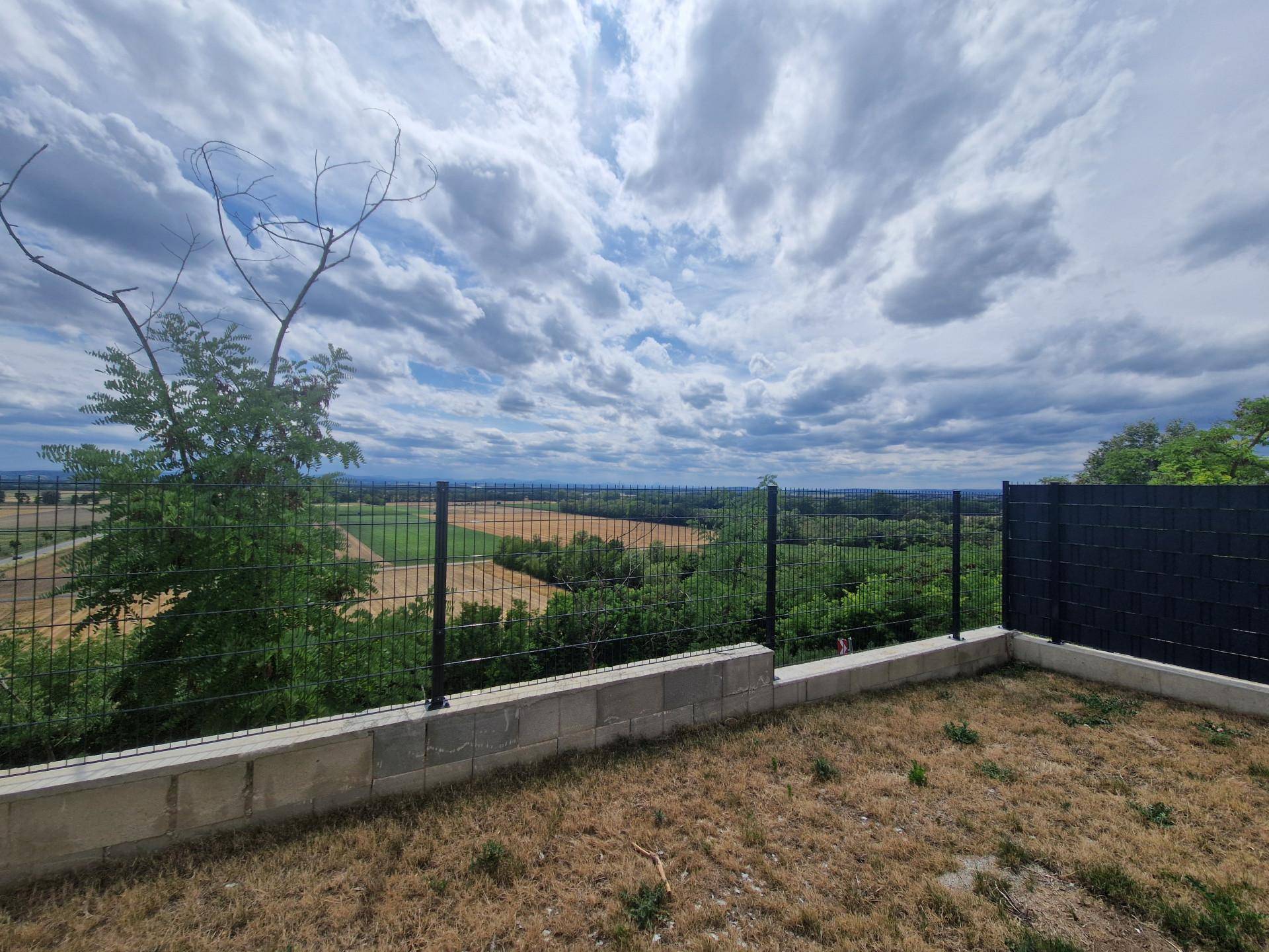 Gartenbereich mit Zaun und weitem Blick über die ländliche Umgebung unter bewölktem Himmel.
