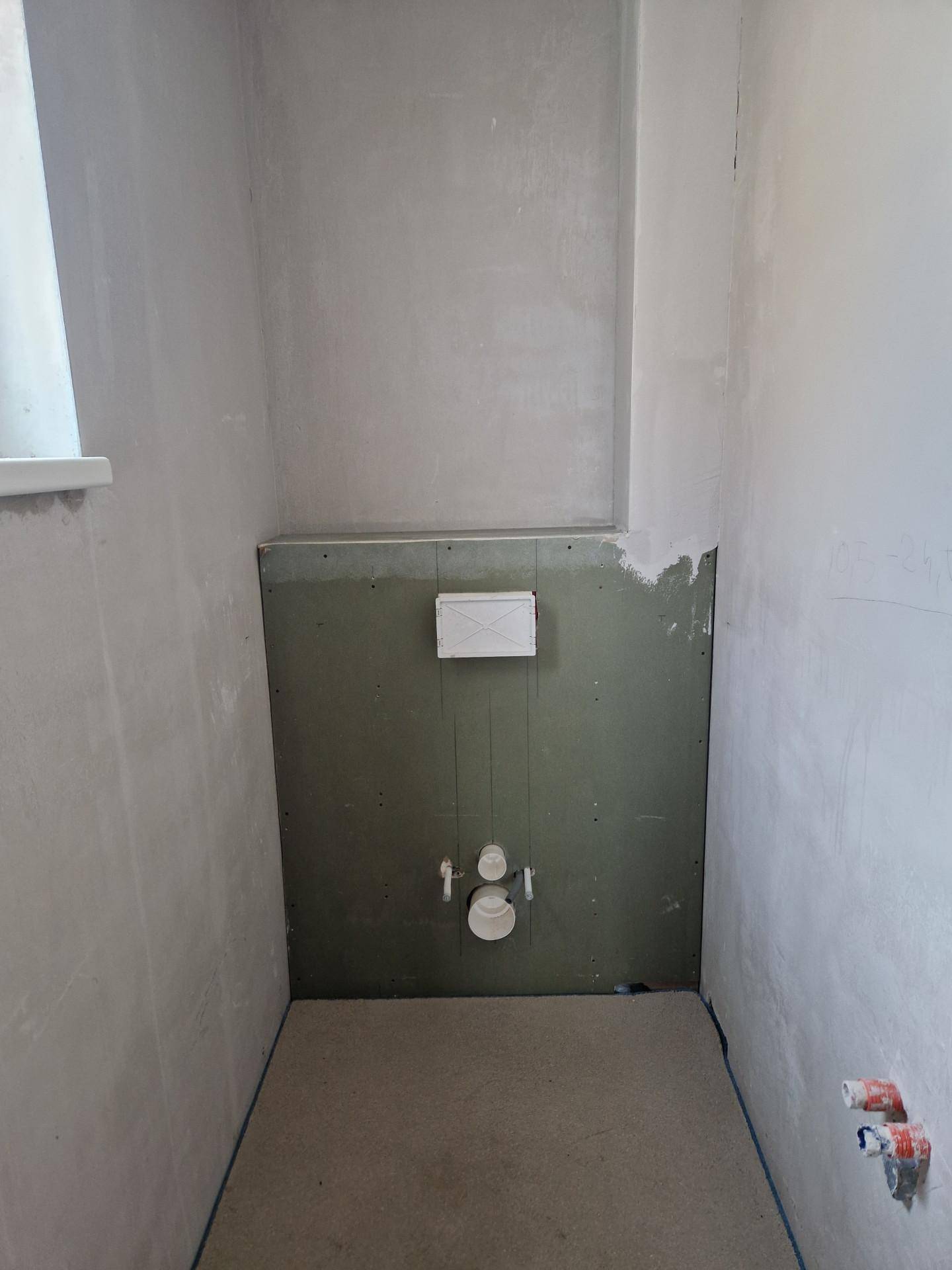 Unfertiger WC-Raum mit vorbereiteten Anschlüssen für eine Wandtoilette und Fenster.