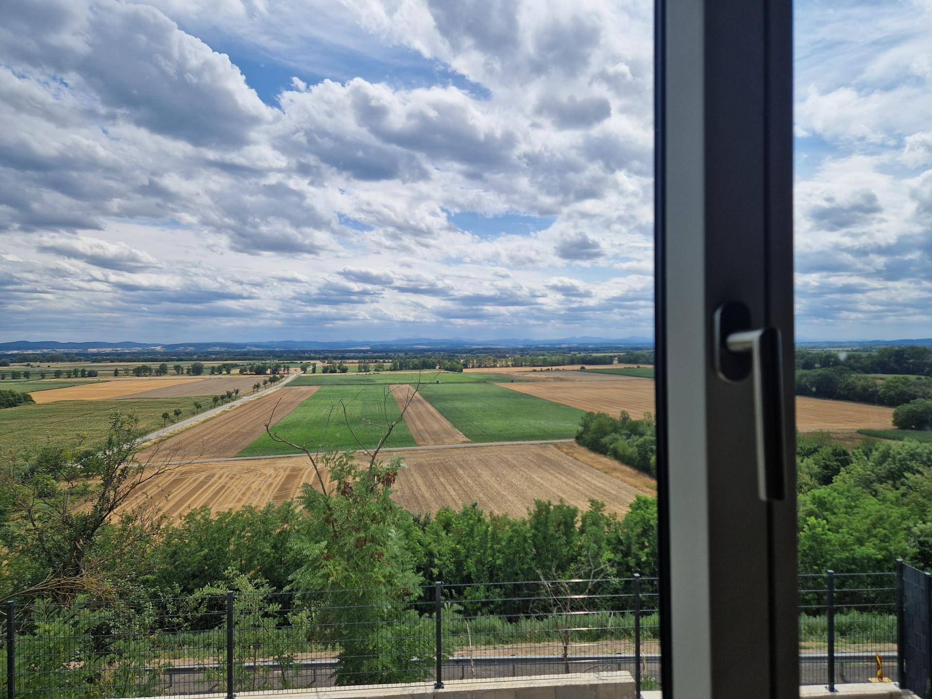 Panoramablick aus dem Fenster auf weite Felder und grüne Hügel unter einem bewölkten Himmel.