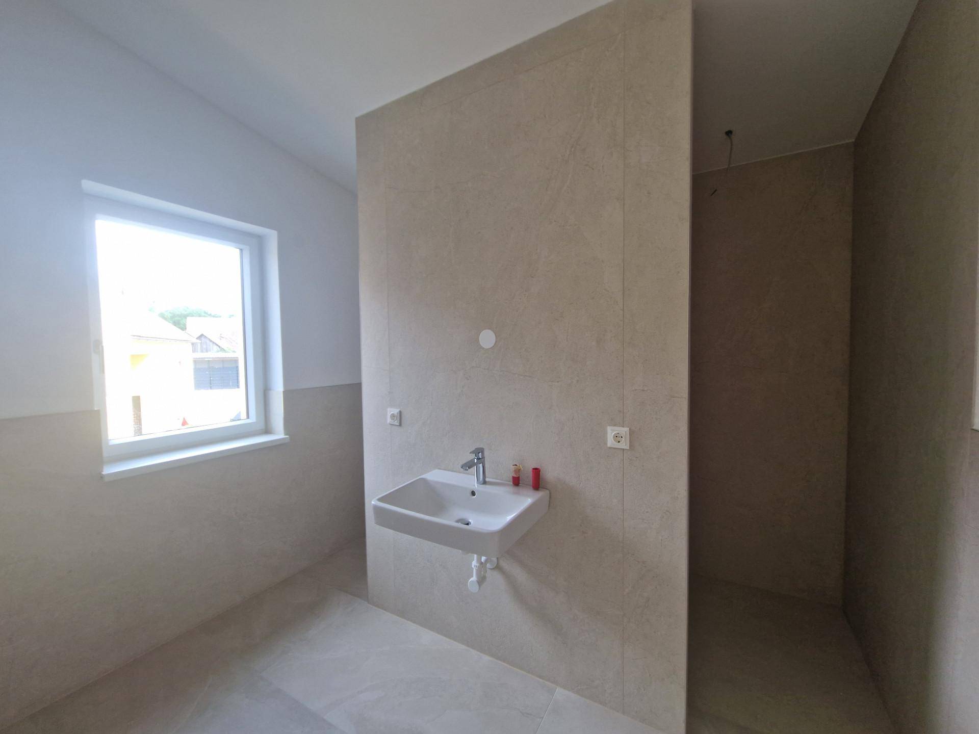 Helles Badezimmer mit Fenster, Waschbecken und vorbereitetem Duschbereich, noch unvollendet.