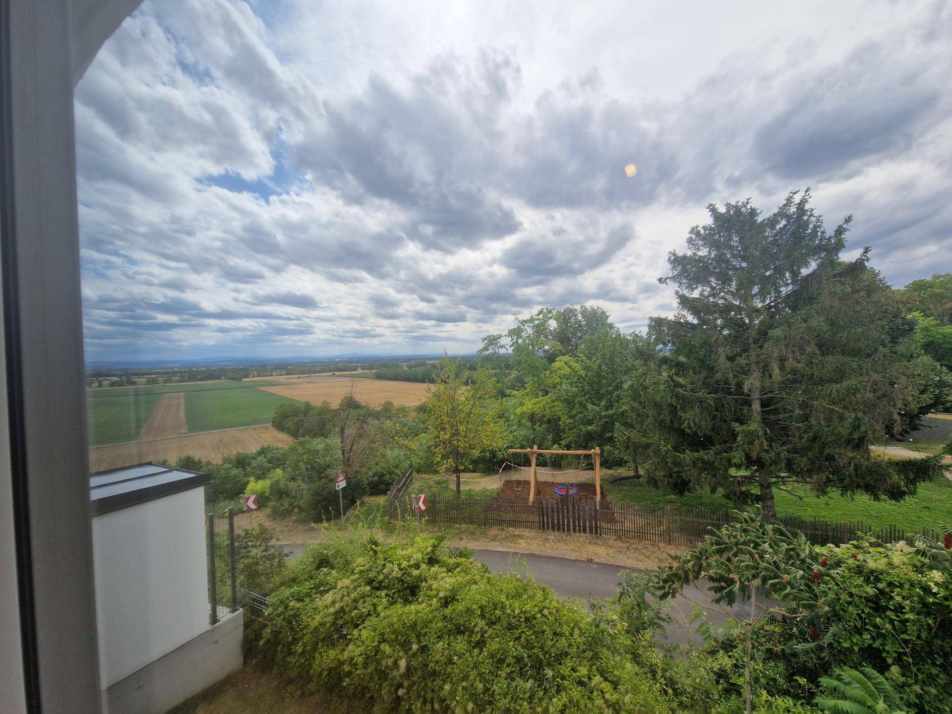 Panoramablick aus dem Fenster auf weite Felder, grüne Bäume und einen Spielbereich.