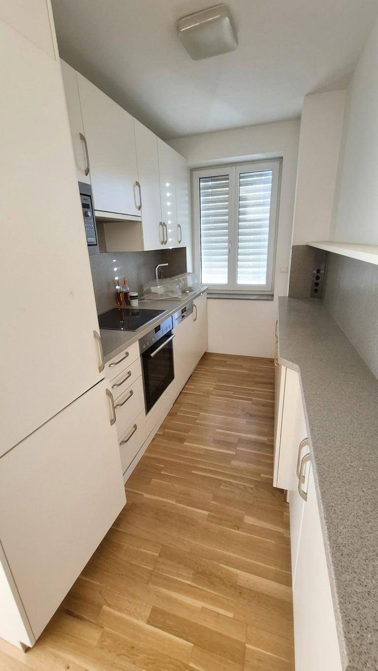 Schmale, moderne Küche mit weißen Schränken, Holzboden und Fenster.