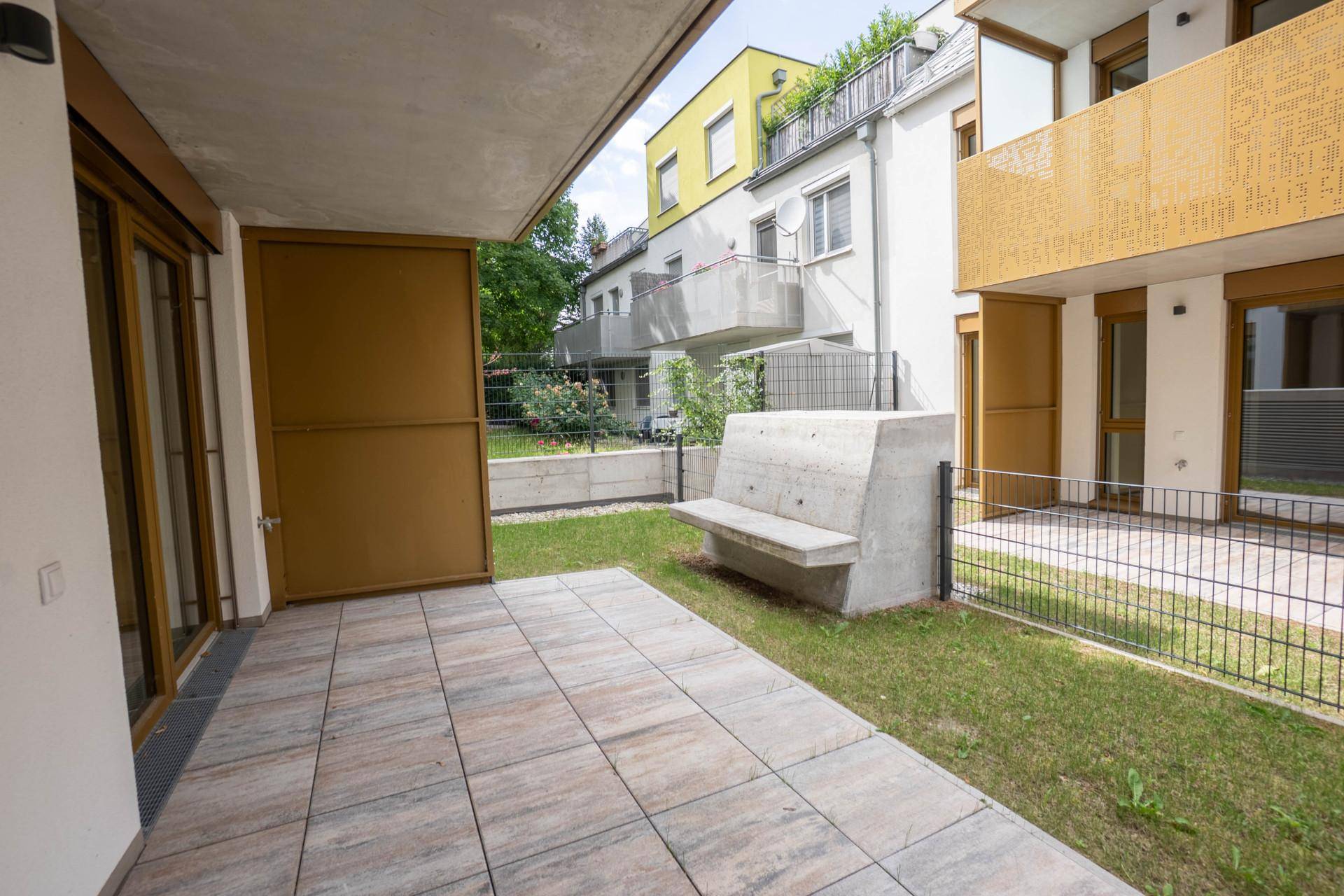 Geflieste Terrasse mit kleiner Rasenfläche und einer modernen Betonbank im Außenbereich.