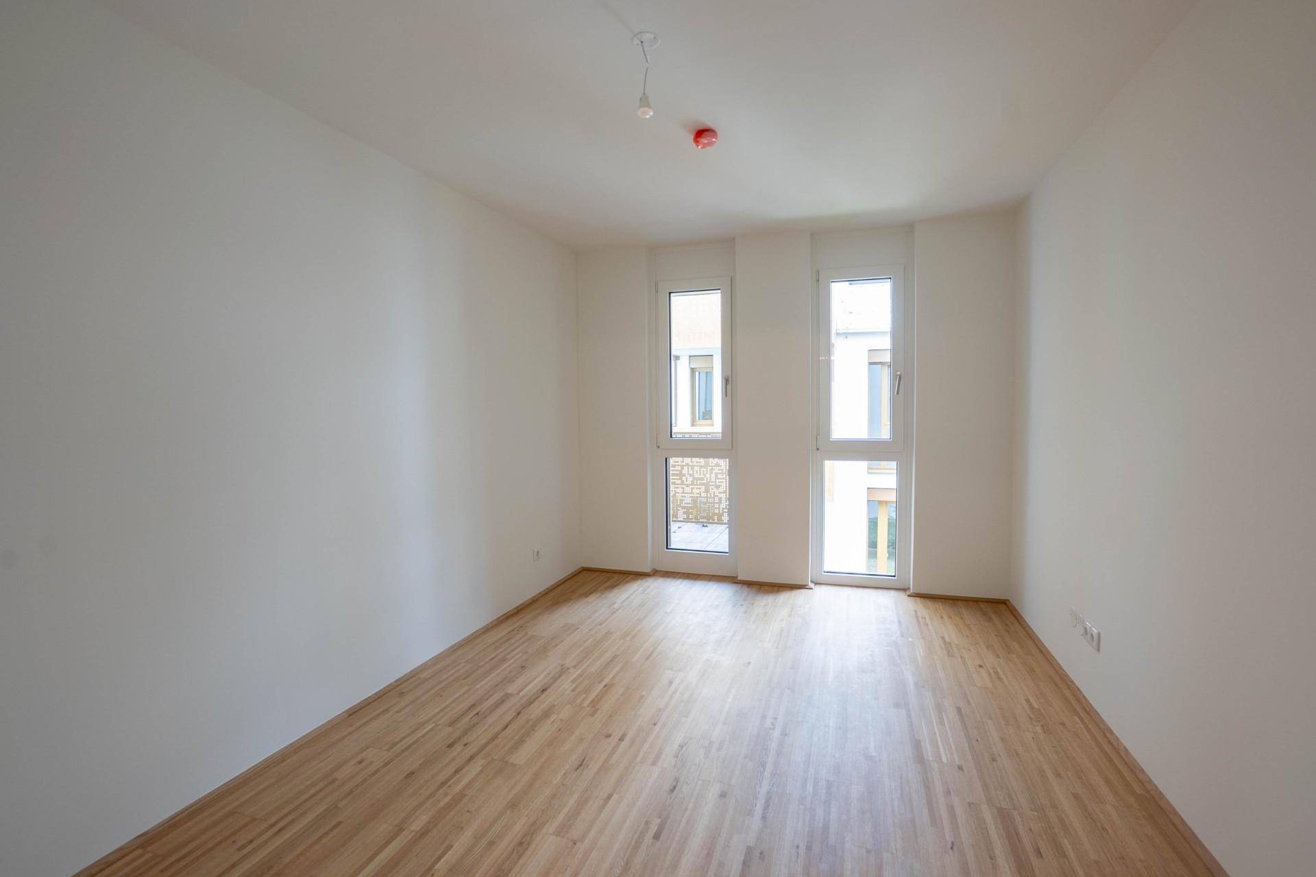 Leeres Schlafzimmer mit Holzboden und zwei Fenstern, eines davon mit Zugang zum Balkon.