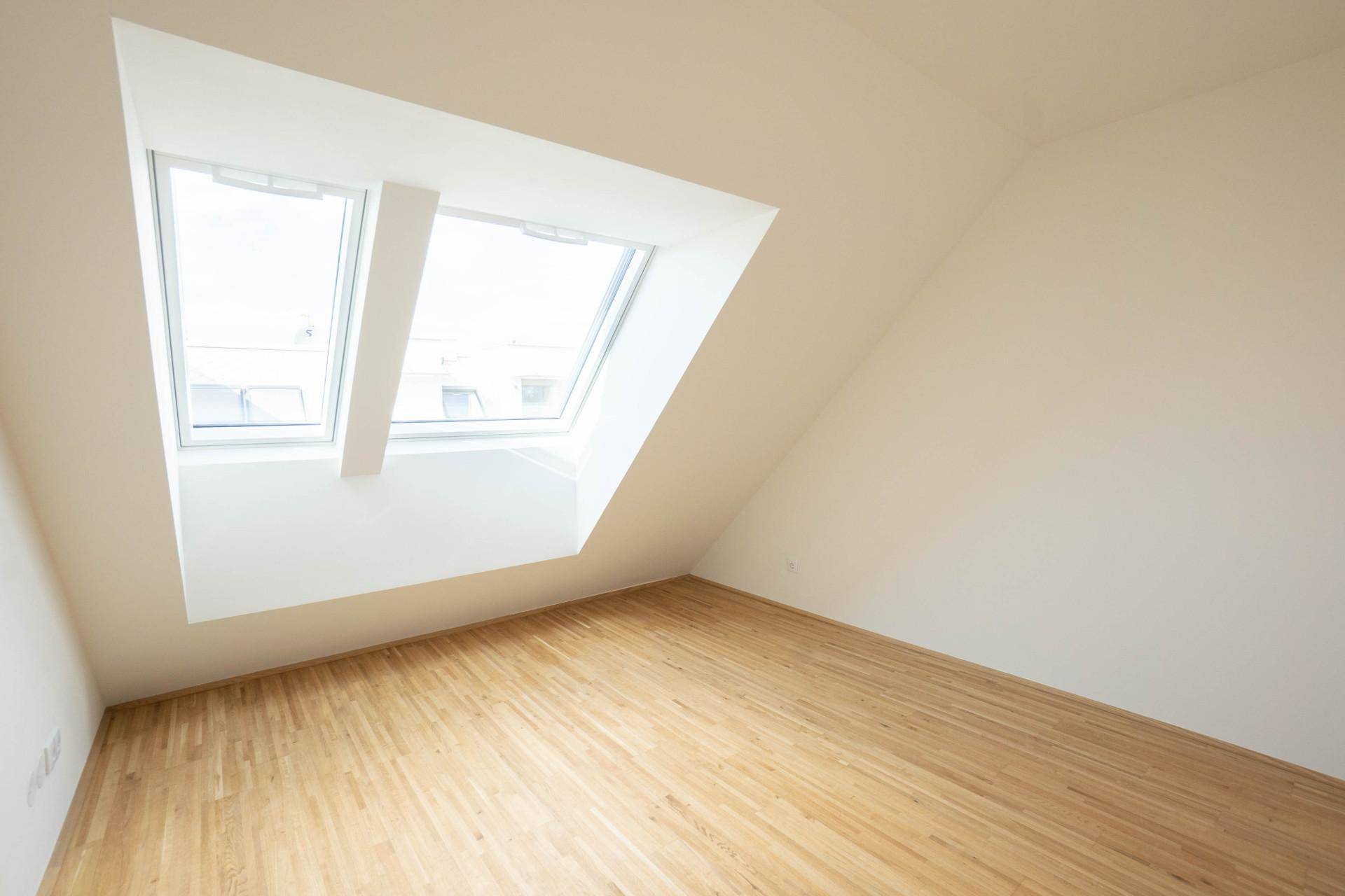 Helles Schlafzimmer im Dachgeschoss mit schrägen Wänden, zwei Dachfenstern und Holzparkettboden.
