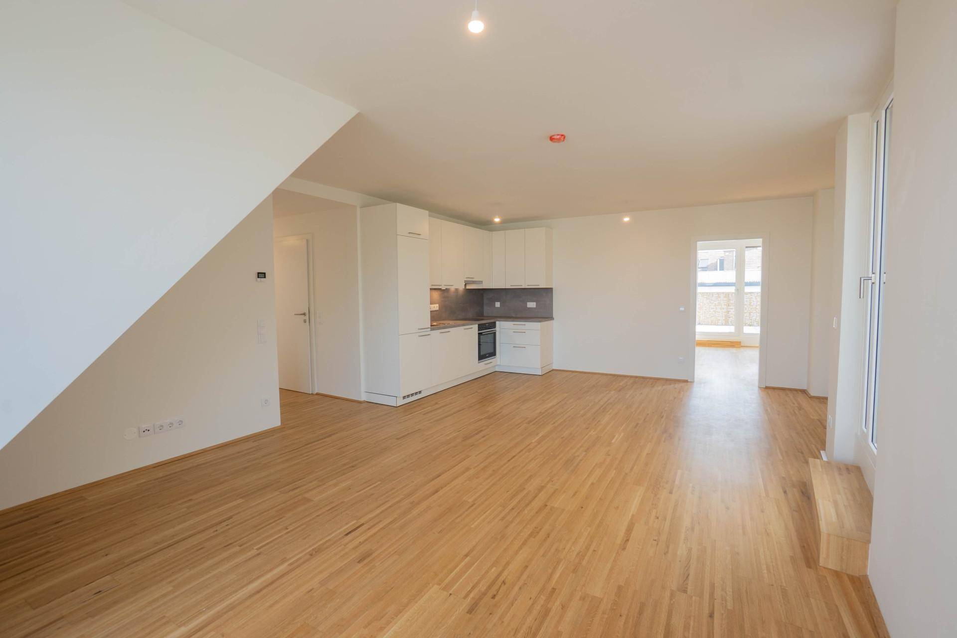 Offener Wohnbereich mit integrierter Küche, hellem Holzfußboden und Zugang zu einem Balkon.