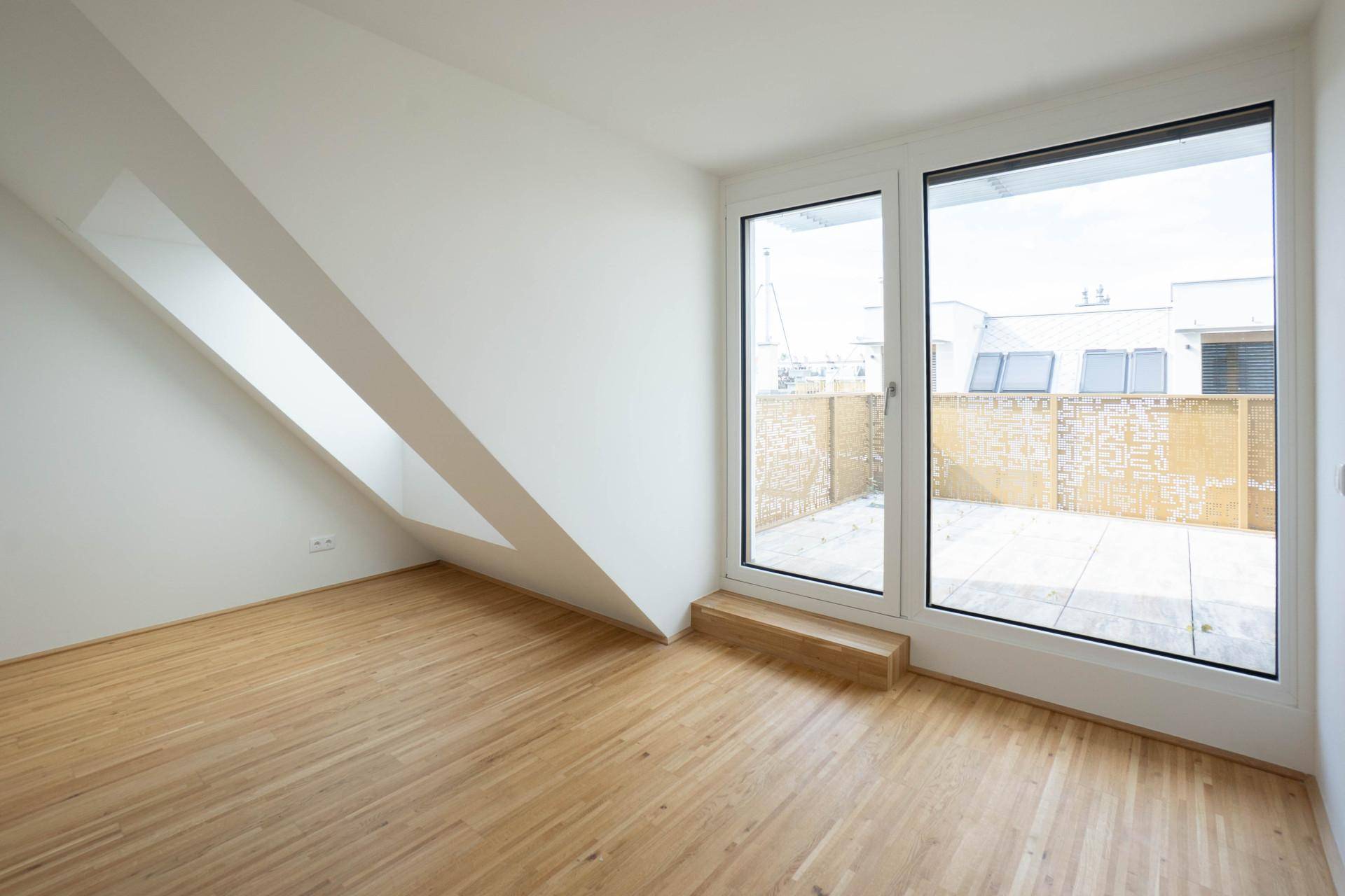 Helles Zimmer mit schräger Wand, großem Fenster und Zugang zu einer Terrasse, ausgestattet mit Holzfußboden.