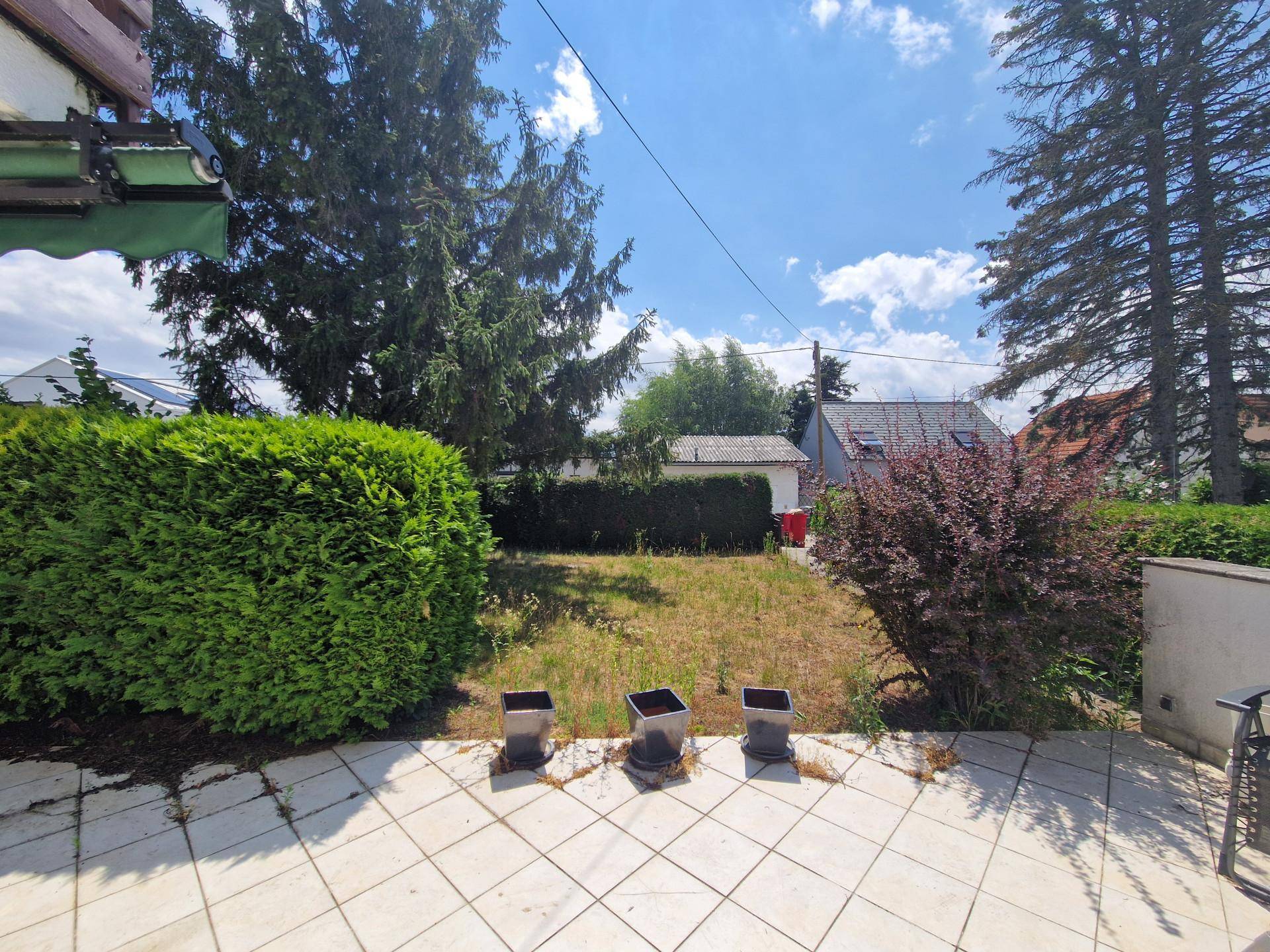 Gartenbereich mit Rasenfläche, Hecken und einer kleinen Terrasse unter blauem Himmel.