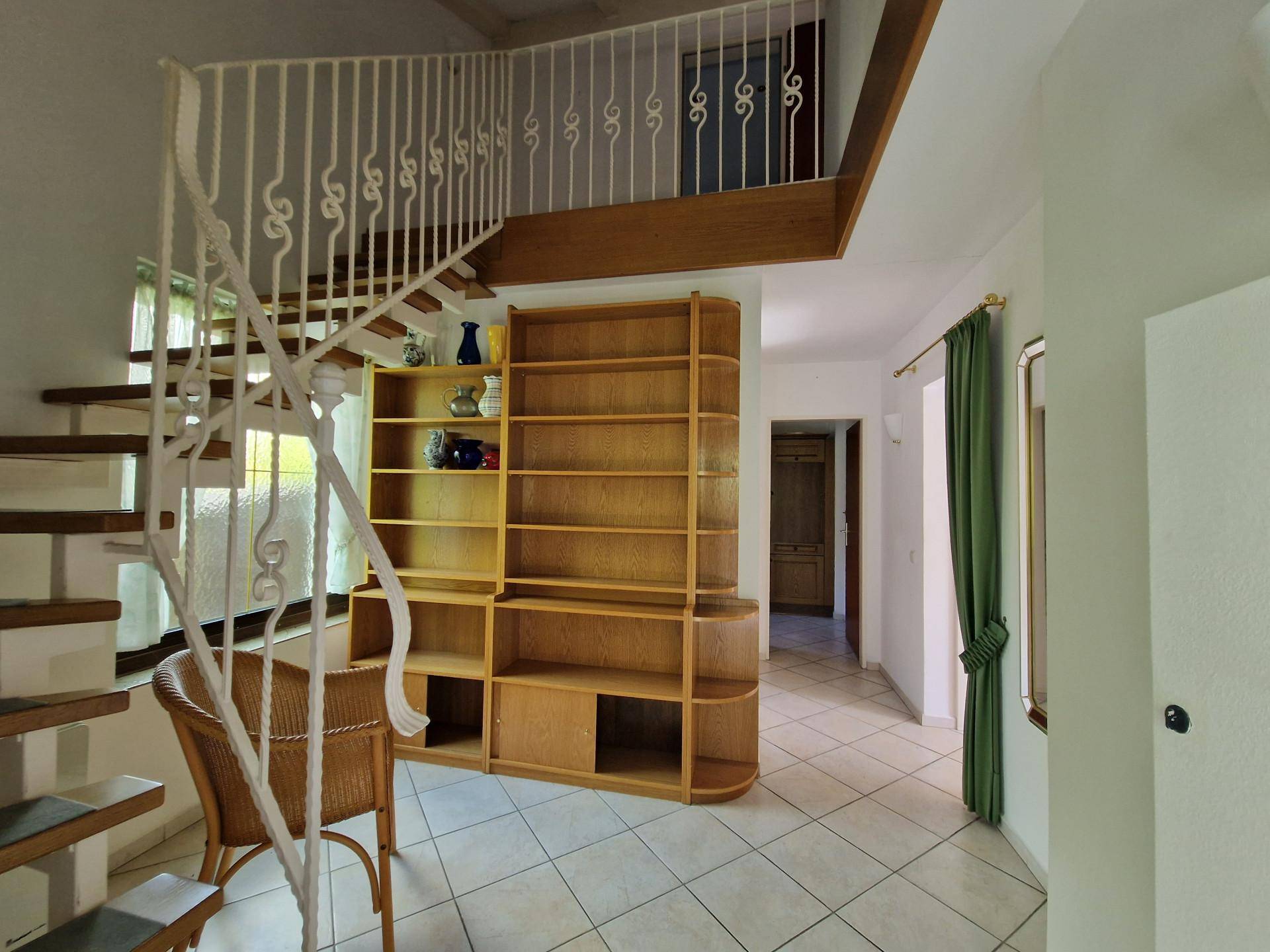 Heller Flur mit geschwungener Treppe, Einbauregal und Fliesenboden.