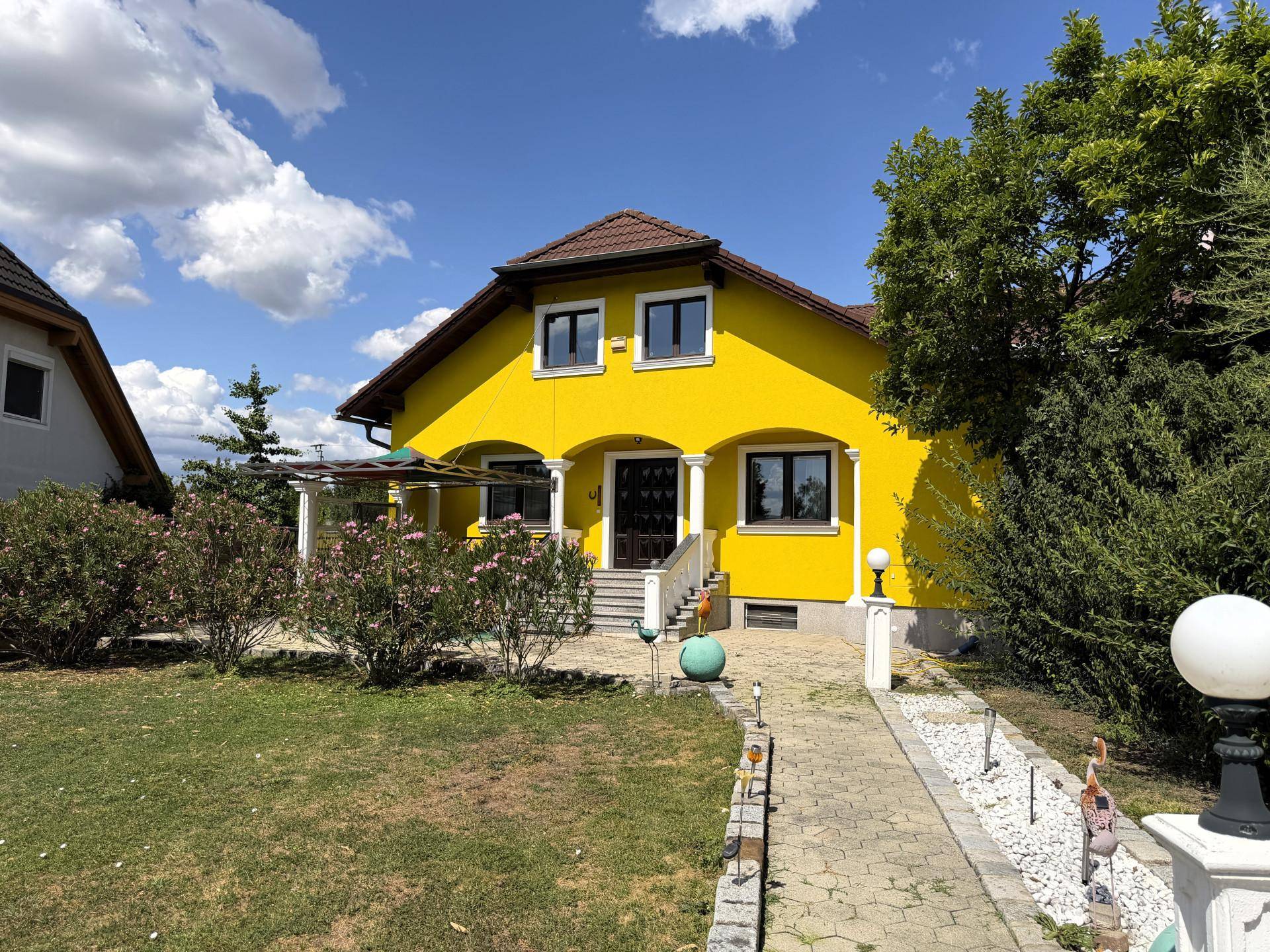Frontansicht des gelben Hauses mit gepflegtem Vorgarten und blühenden Sträuchern unter blauem Himmel.