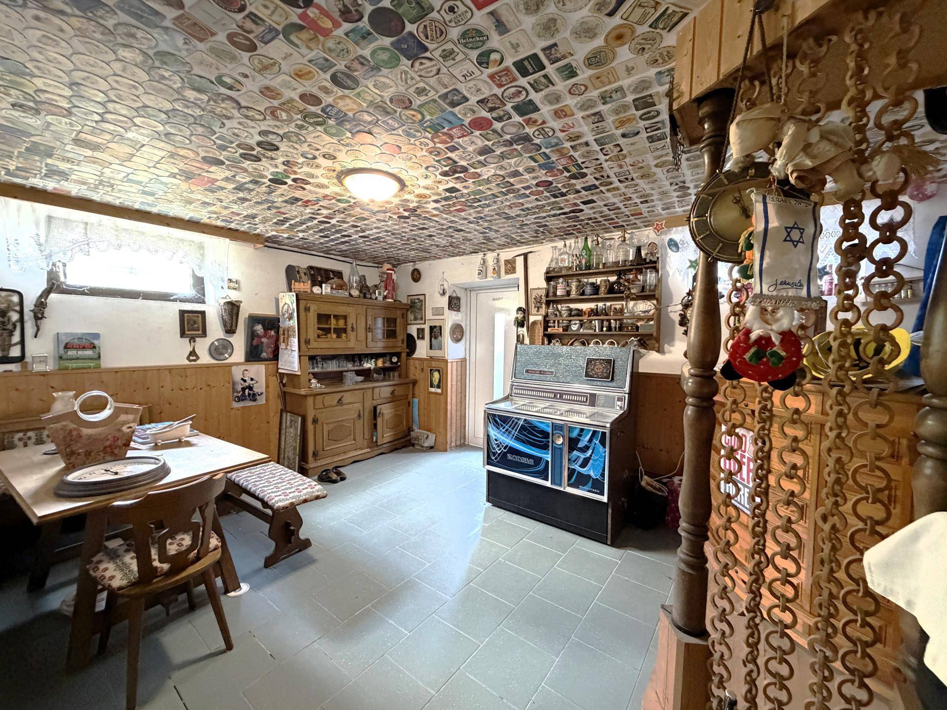 Rustikaler Partykeller mit Jukebox, Holztisch und einer Decke voller Bierdeckel.
