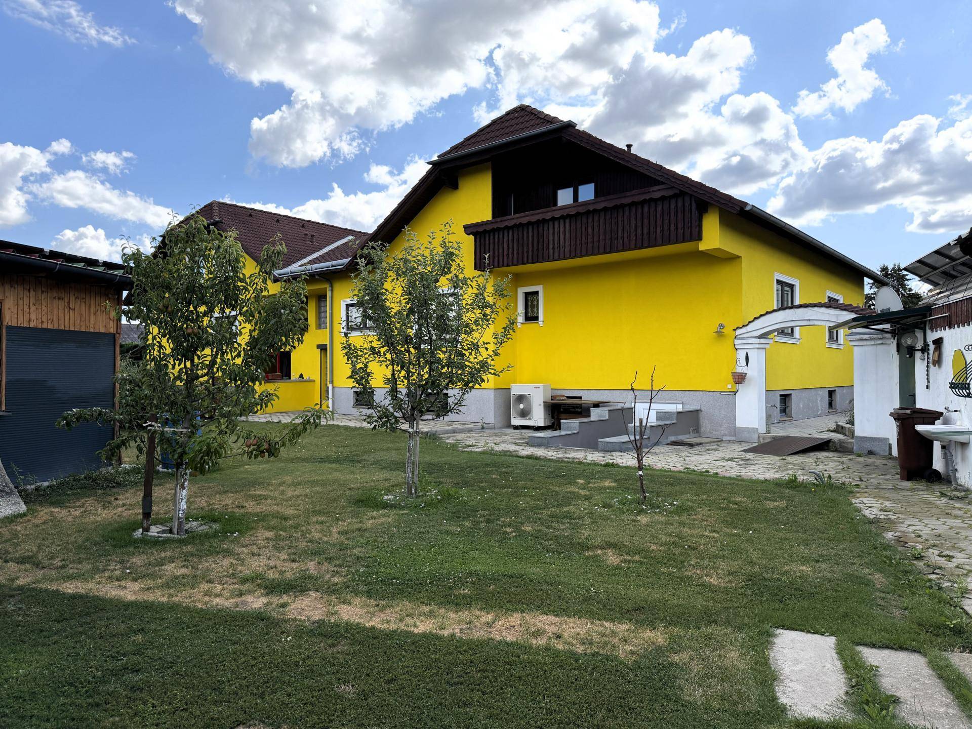 Rückansicht des gelben Hauses mit Garten, jungen Bäumen und einer Außeneinheit der Klimaanlage.