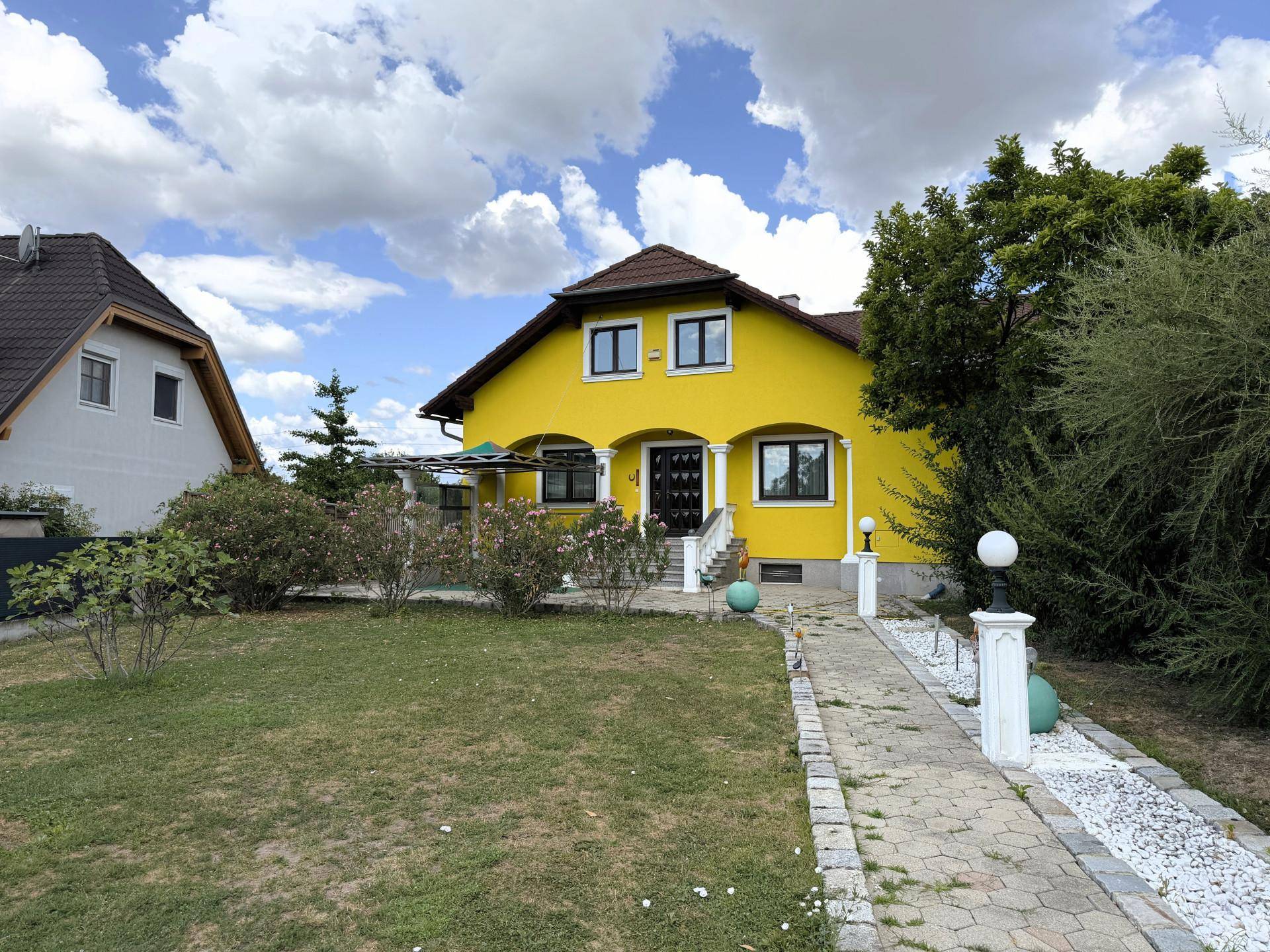 Frontansicht des gelben Hauses mit Vorgarten und gepflastertem Weg unter blauem Himmel.