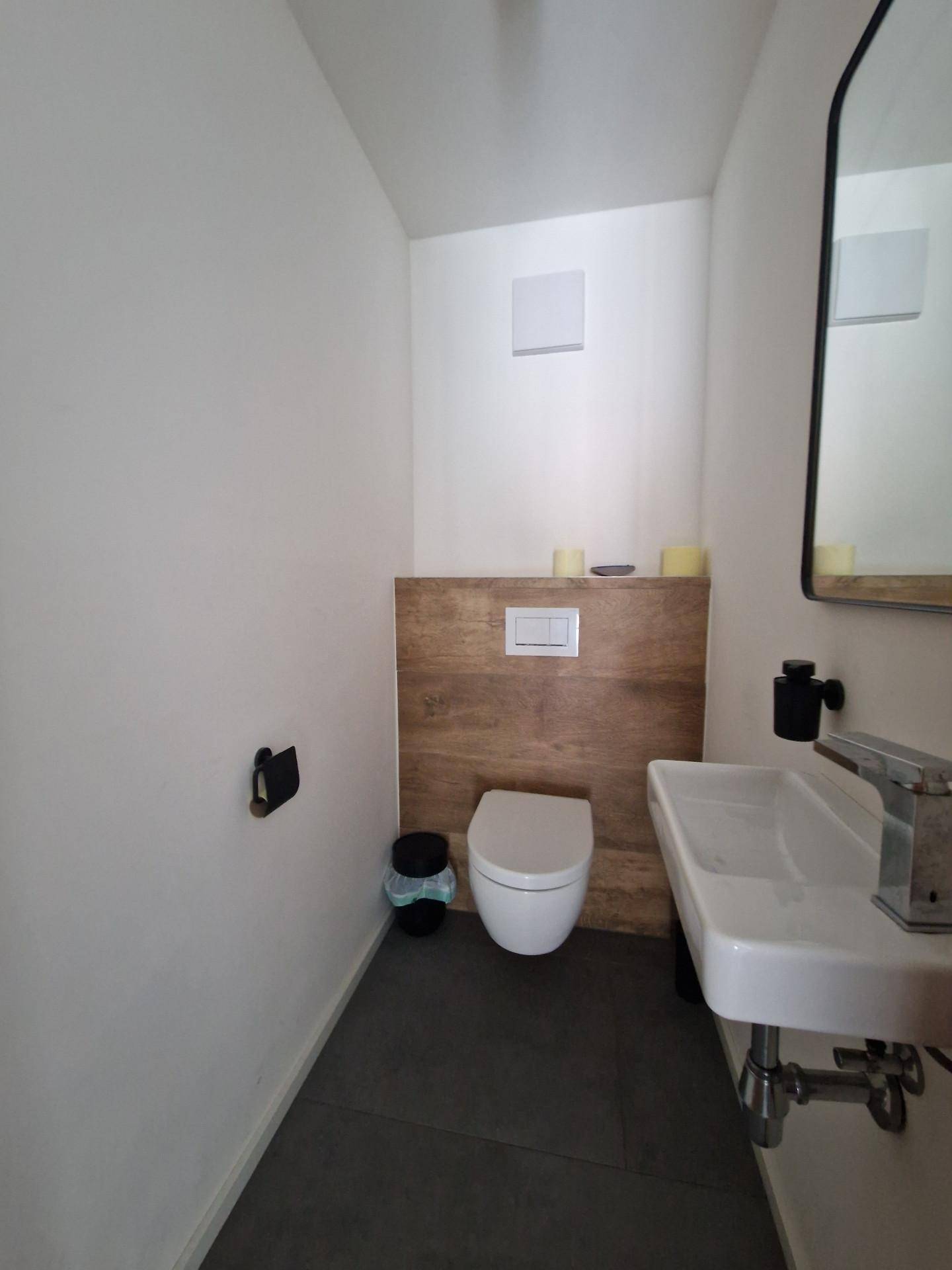 Modernes Gäste-WC mit dunklen Fliesen, wandhängendem WC und einem Waschbecken mit Holzverkleidung.