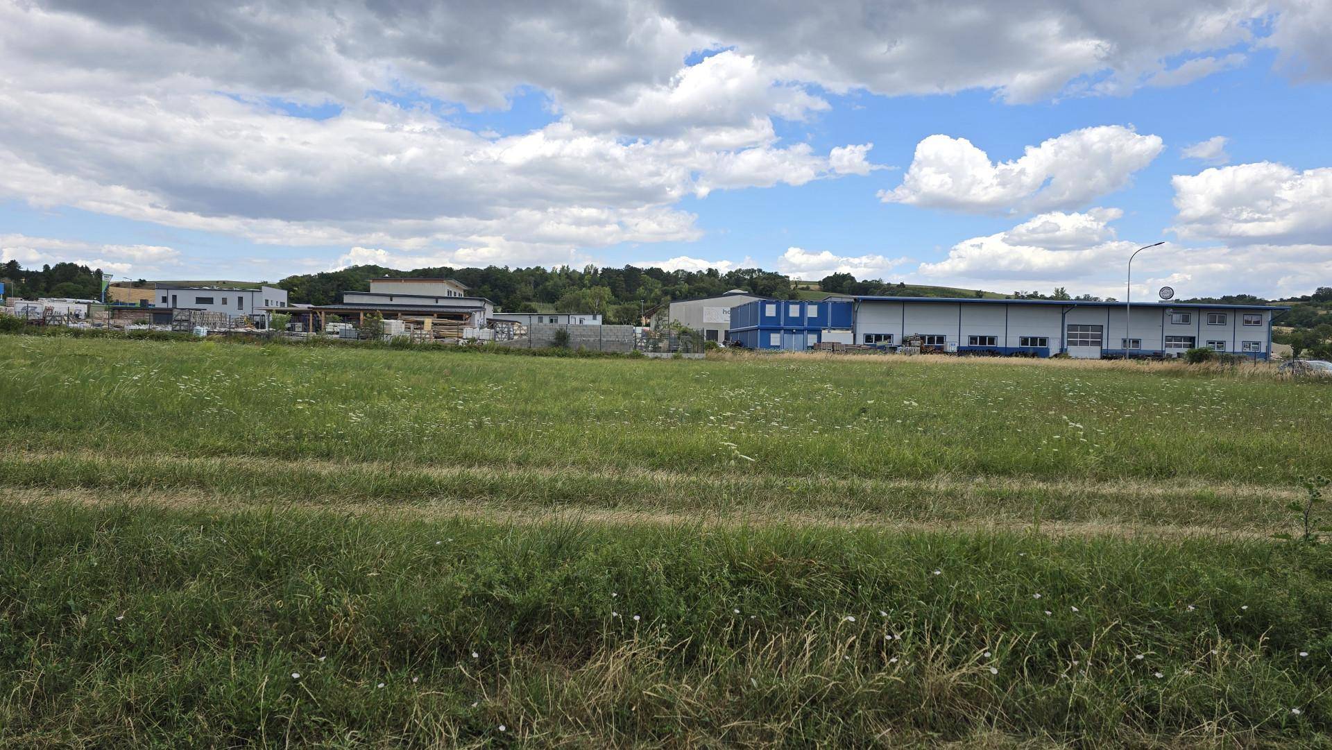 Weitläufiges grünes Feld mit verschiedenen Industrie- und Gewerbegebäuden im Hintergrund unter einem teilweise bewölkten Himmel.