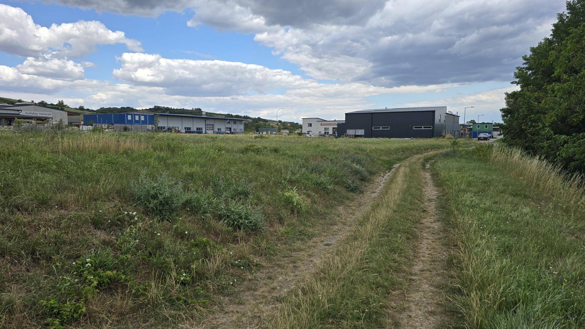 Ein Feldweg schlängelt sich durch eine grüne Wiese mit Gewerbegebäuden und Bäumen im Hintergrund unter bewölktem Himmel.