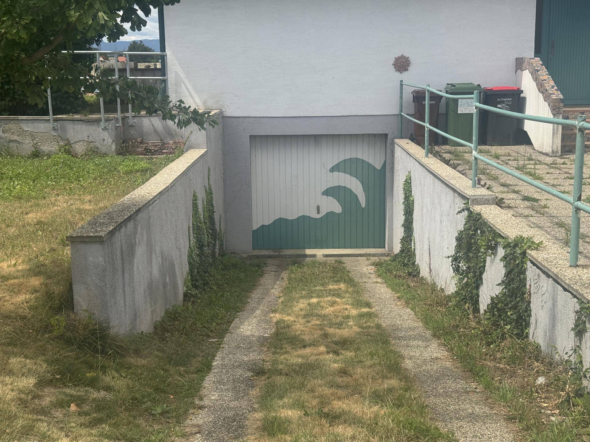 Einfahrt zu einer Tiefgarage mit einer kreativ bemalten Tür, umgeben von Betonmauern und Grün.