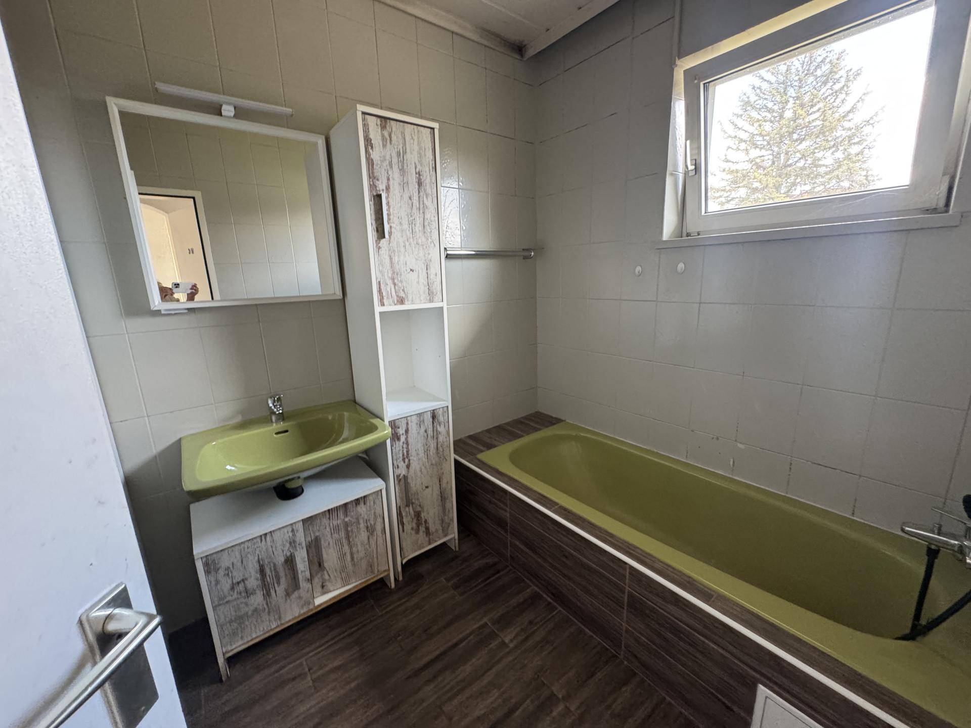Badezimmer mit grüner Badewanne und Waschbecken, umgeben von hellen Fliesen und einem Fenster.