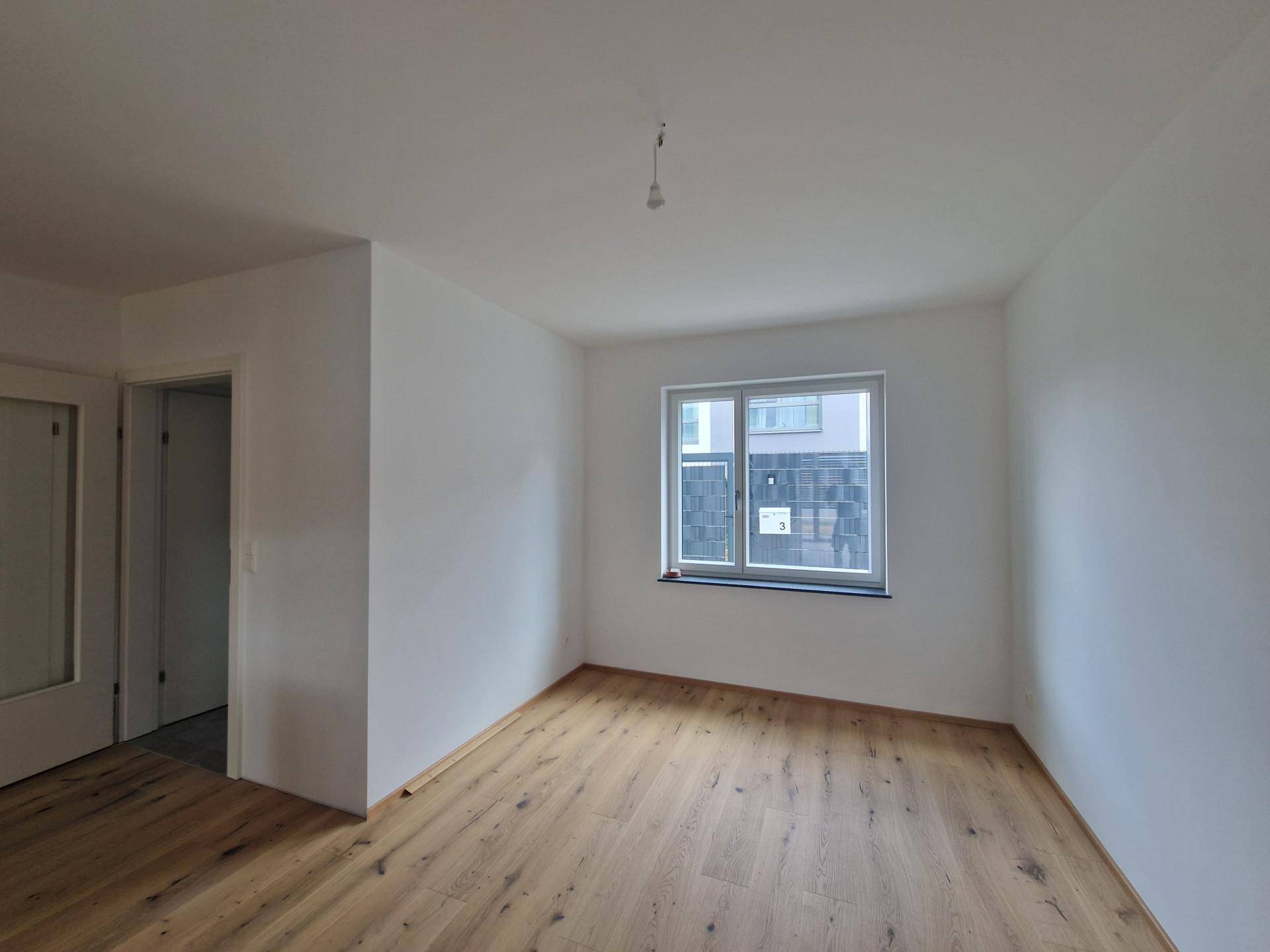 Leeres Zimmer mit hellem Holzboden und einem Fenster, das Tageslicht hereinlässt.