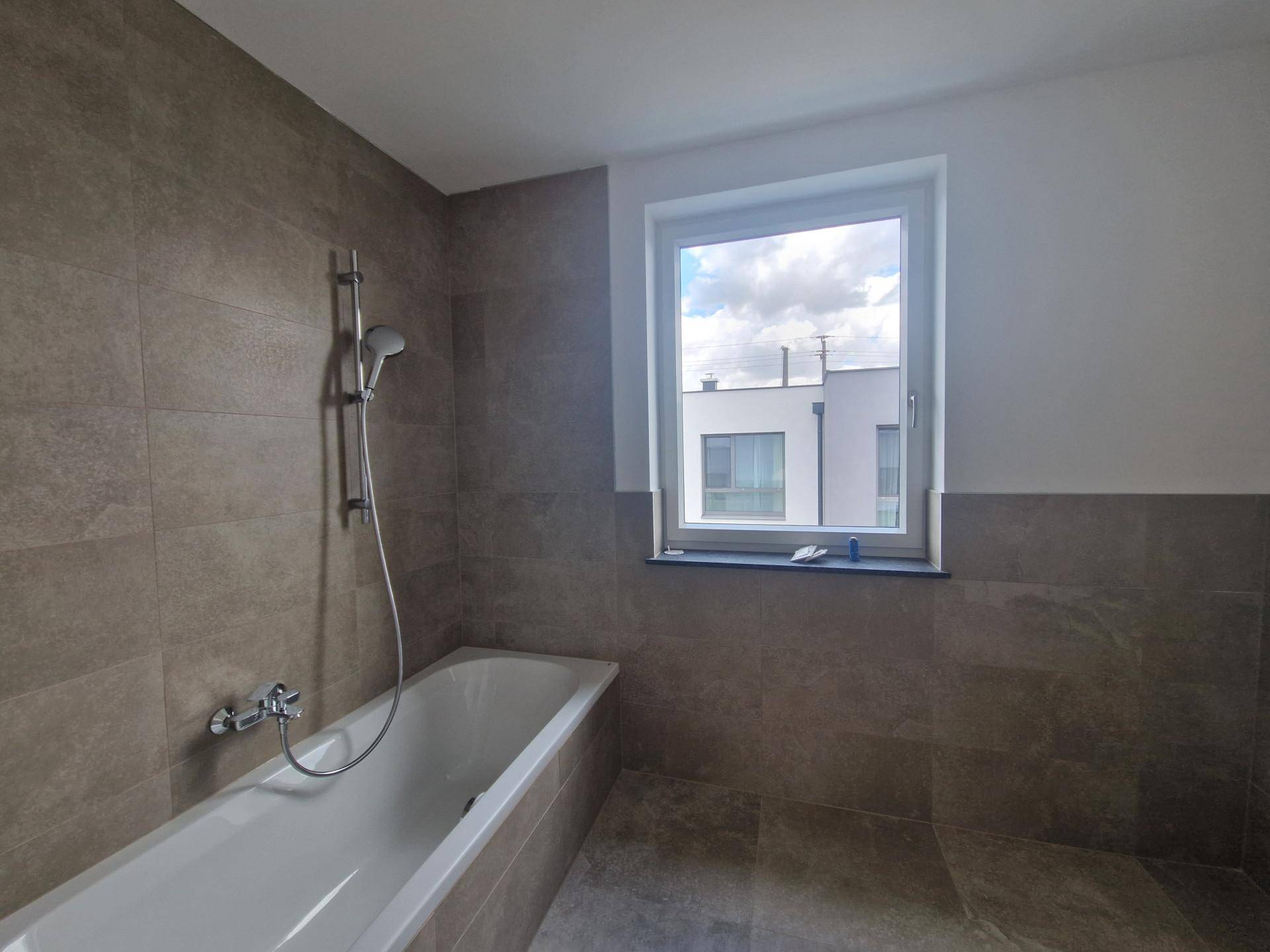 Badezimmer mit Badewanne, grauen Wandfliesen und einem Fenster für natürliches Licht.