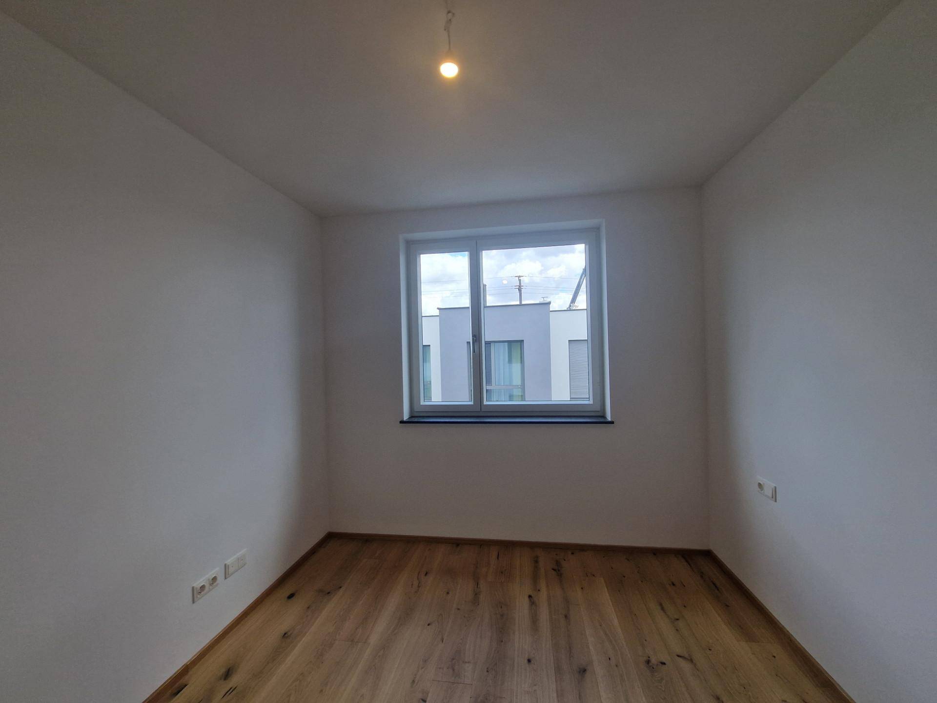Helles Schlafzimmer mit Holzboden und einem Fenster, das den Blick auf Nachbargebäude freigibt.