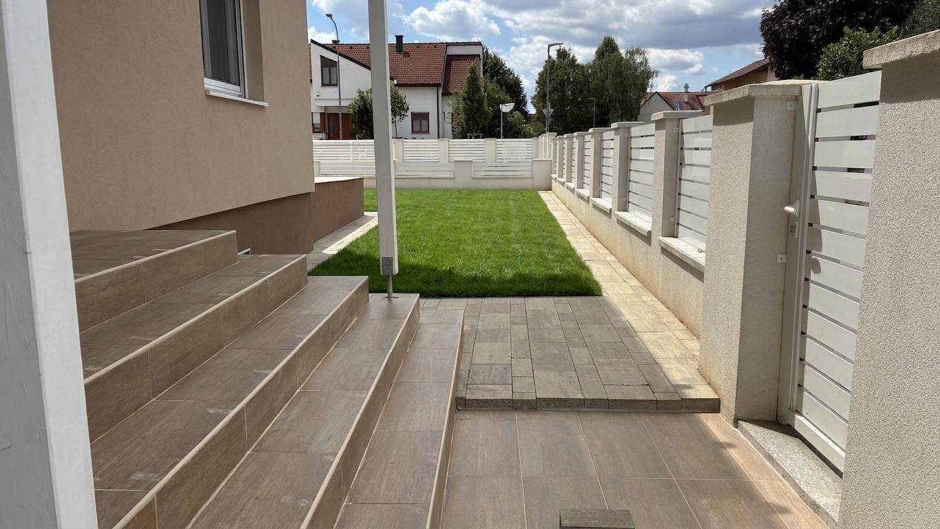 Außenbereich mit Steintreppe, gepflastertem Weg und Rasenfläche, umgeben von einem modernen Zaun.
